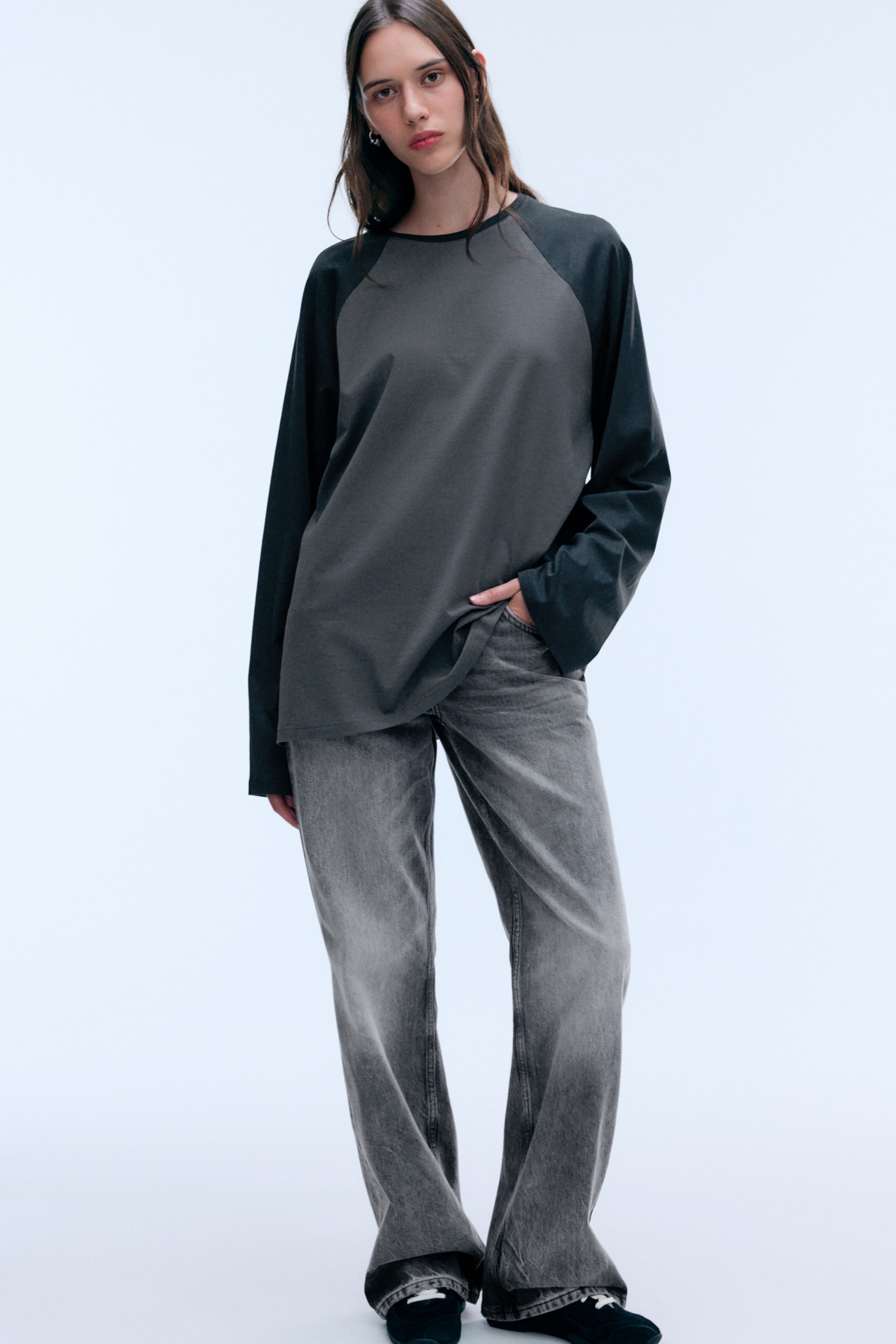 Vezi imaginea mai mare: Un top lejer cu mâneci lungi raglan, având corpul de culoare gri mediu și mâneci contrastante gri închis, este purtat de model. Modelul asortează topul cu o pereche de blugi gri decolorați, sprijinindu-și o mână într-un buzunar, ceea ce lasă tivul cămășii să cadă ușor.