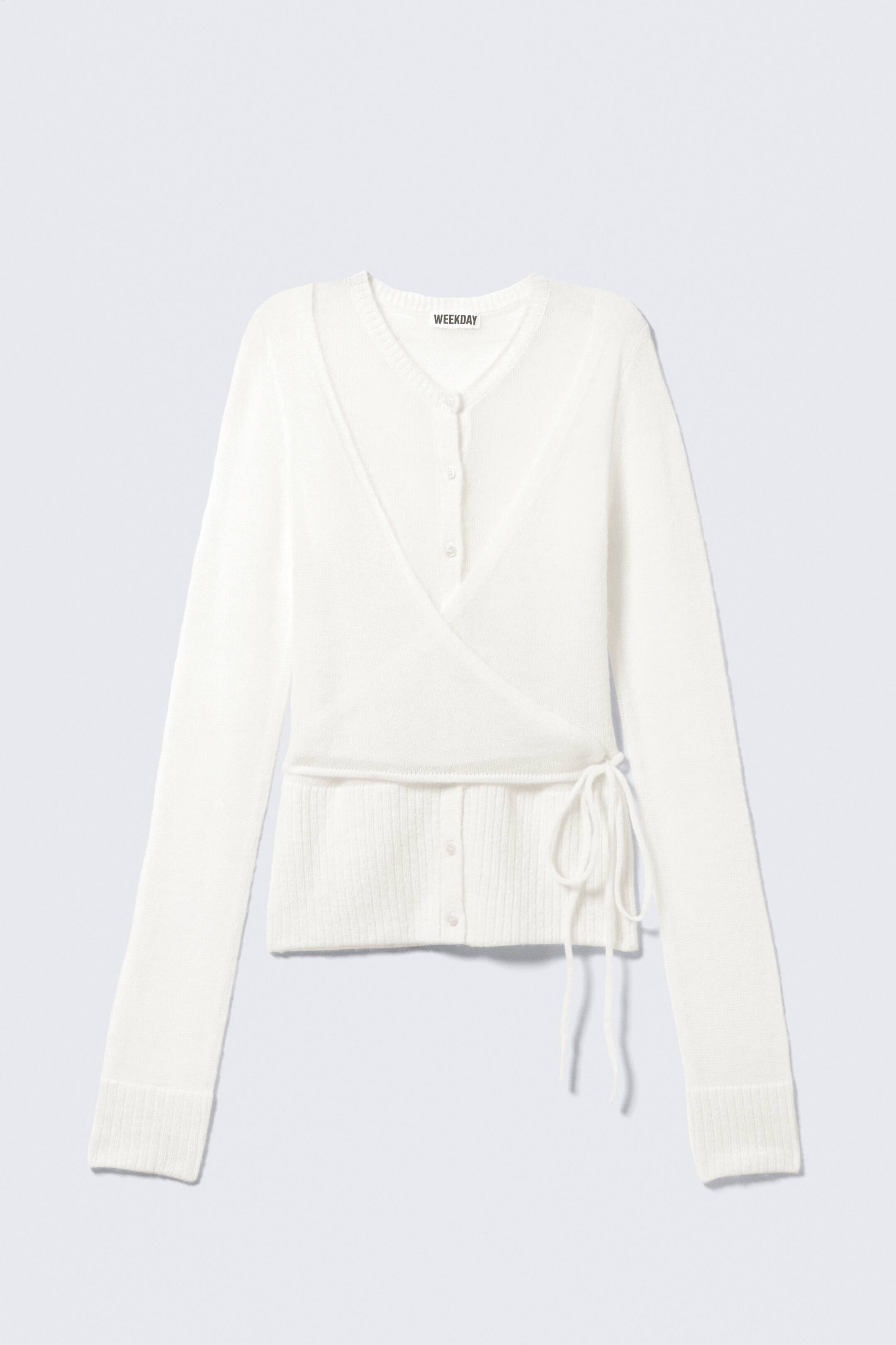 View larger image: A white wrap cardigan is showcased from the front, featuring a V-neckline and a wrap-style front secured with a tie closure on the left side of the waist. Buttons are visible beneath the wrap, and the long sleeves end in ribbed cuffs, while the bottom hem is also ribbed.