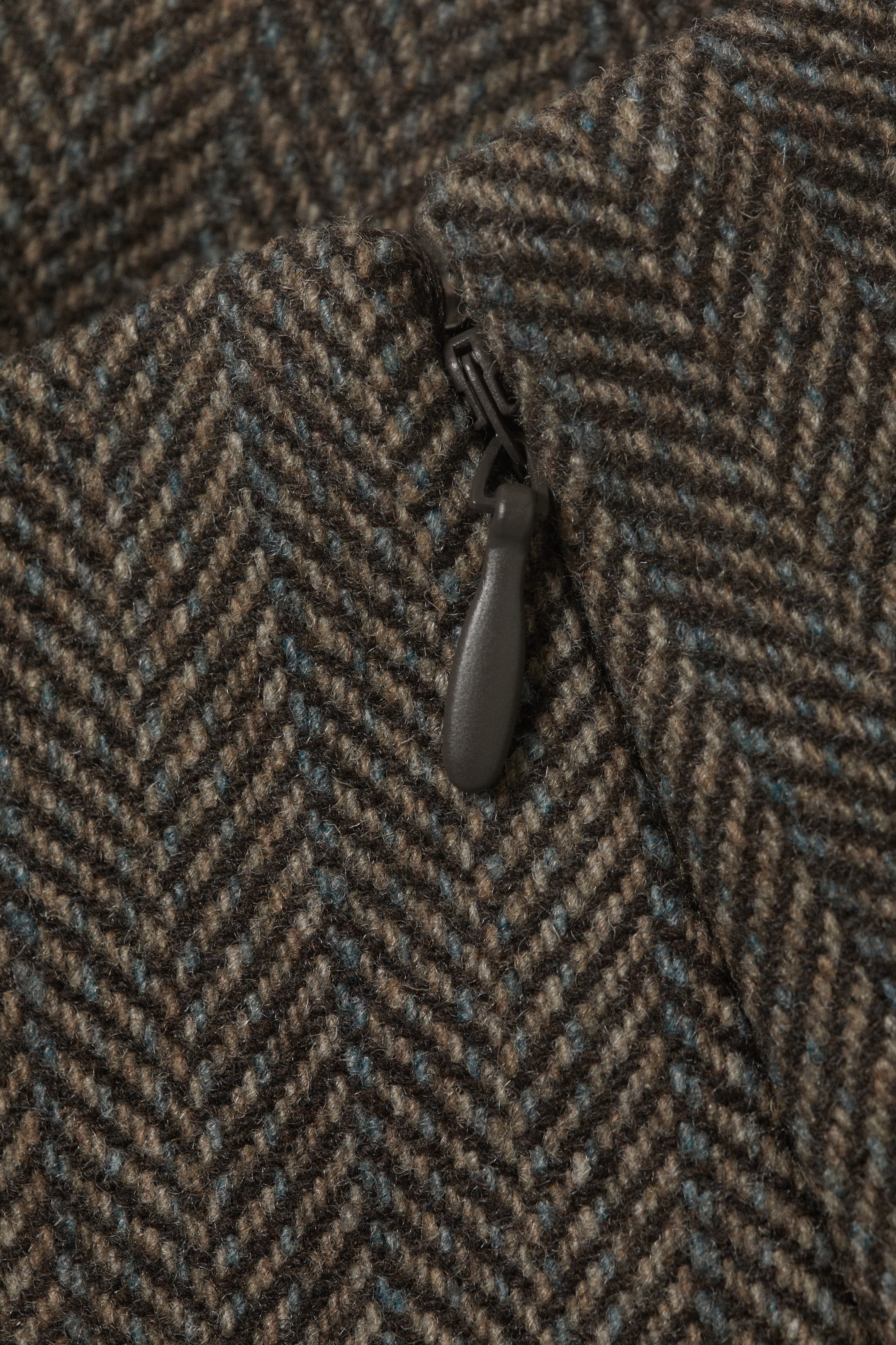 View larger image: A brown and tan herringbone fabric with subtle blue flecks is displaying a dark grey coil zipper. The zipper's plastic pull tab is resting on the fabric surface.