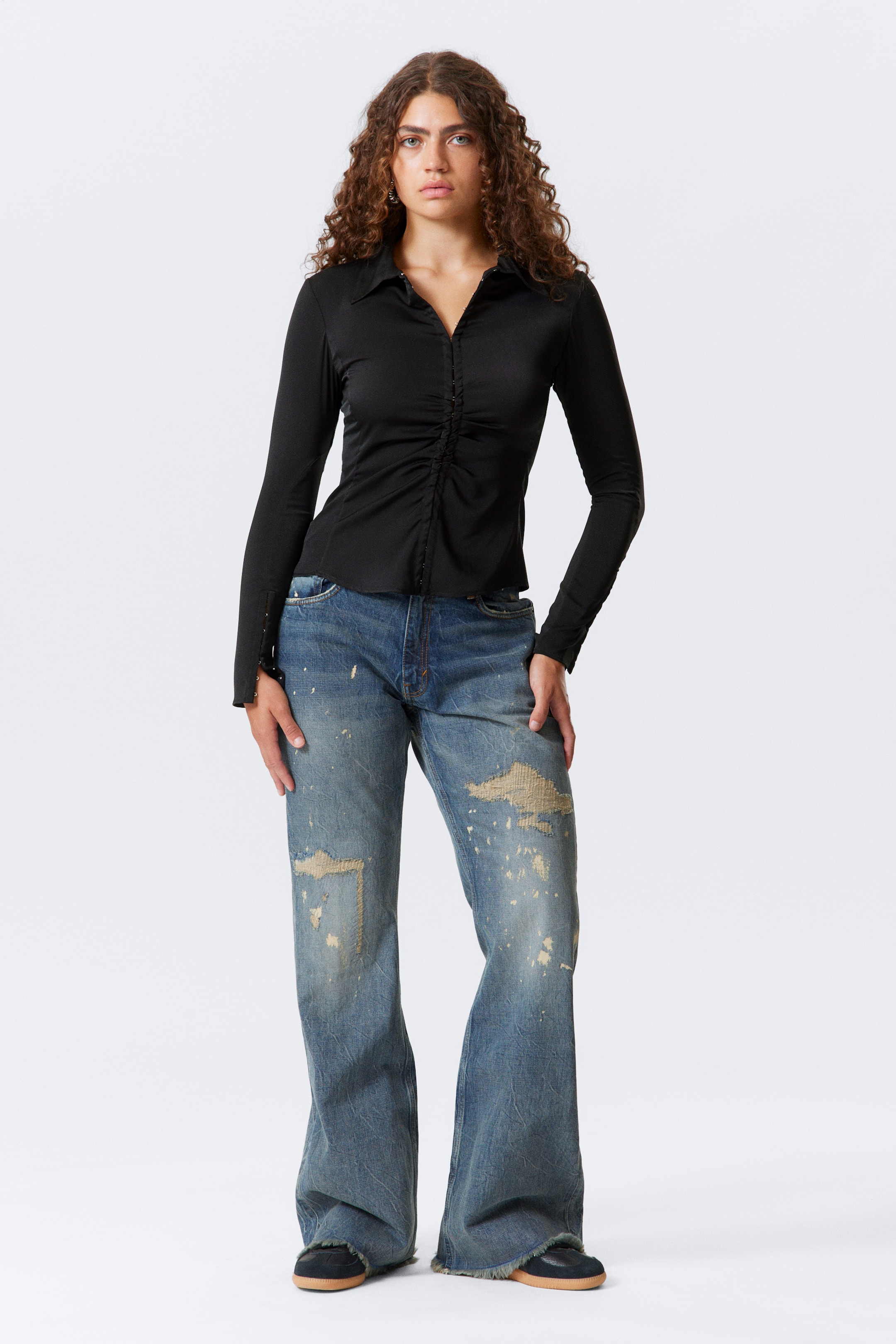 Se større billede: En sort langærmet top, lavet af et glat strikket stof, har en spids krave og en central rynket detalje med små hægter og øjer, båret over slidte blå jeans med brede ben. Jeansene har en falmet vask, synlige lapper og flossede kanter, med en afslappet pasform.