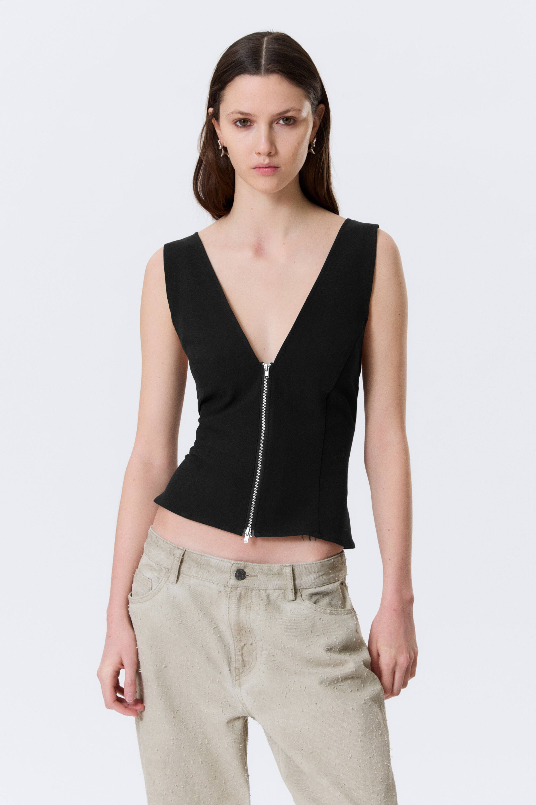 Se større billede: En sort ærmeløs top med en dyb V-udskæring og en sølvfarvet frontlynlås, der er delvist lynet op. Toppen har en tætsiddende silhuet med en kort kant.