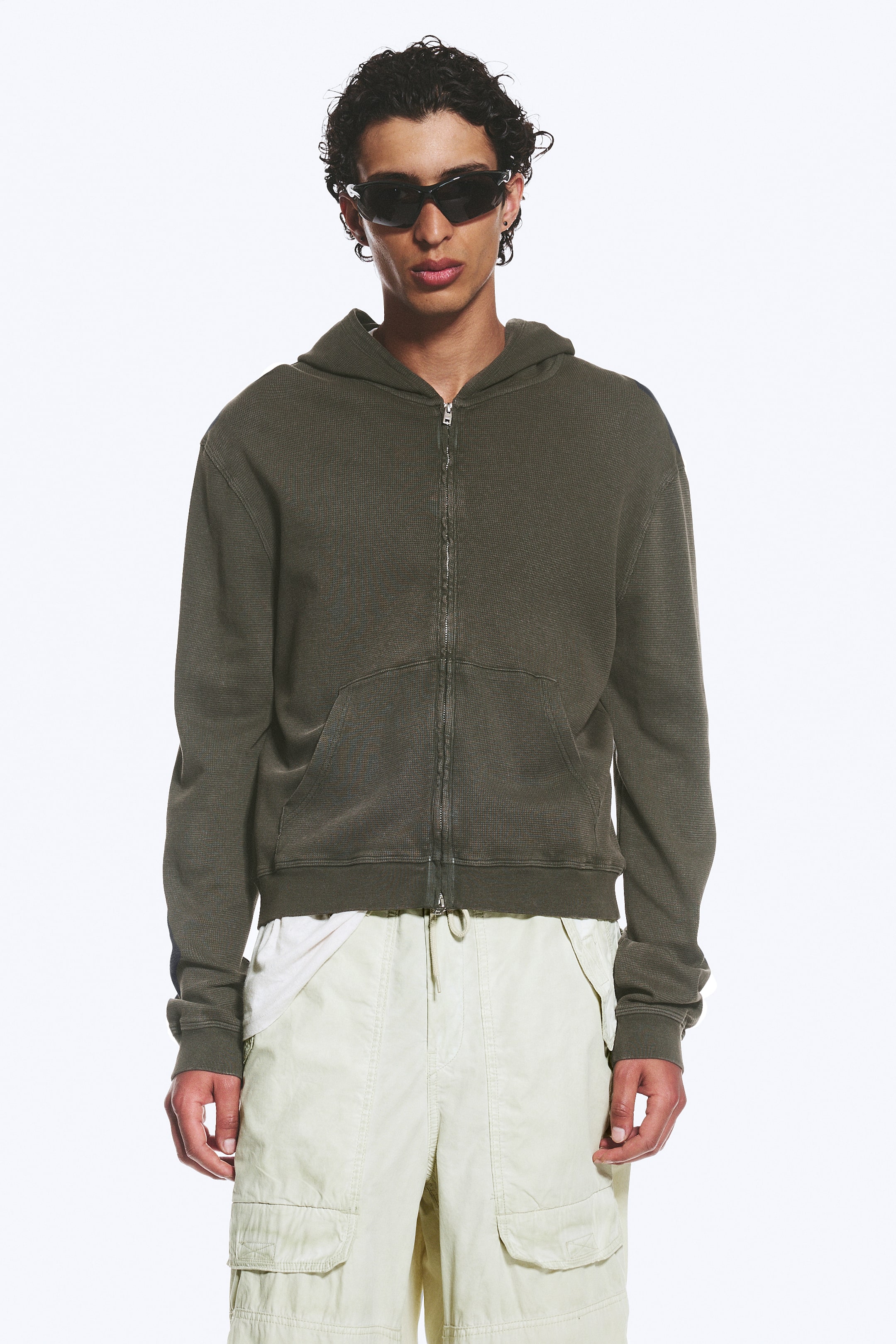 Agrandir l'image: Un sweat à capuche zippé intégral vert olive foncé est présenté de face, mettant en avant un tissu en maille gaufrée et une coupe décontractée. Il est doté d'une capuche attachée, d'une fermeture éclair intégrale, d'une poche kangourou, de poignets côtelés et d'un ourlet côtelé usé, avec un t-shirt blanc en dessous.