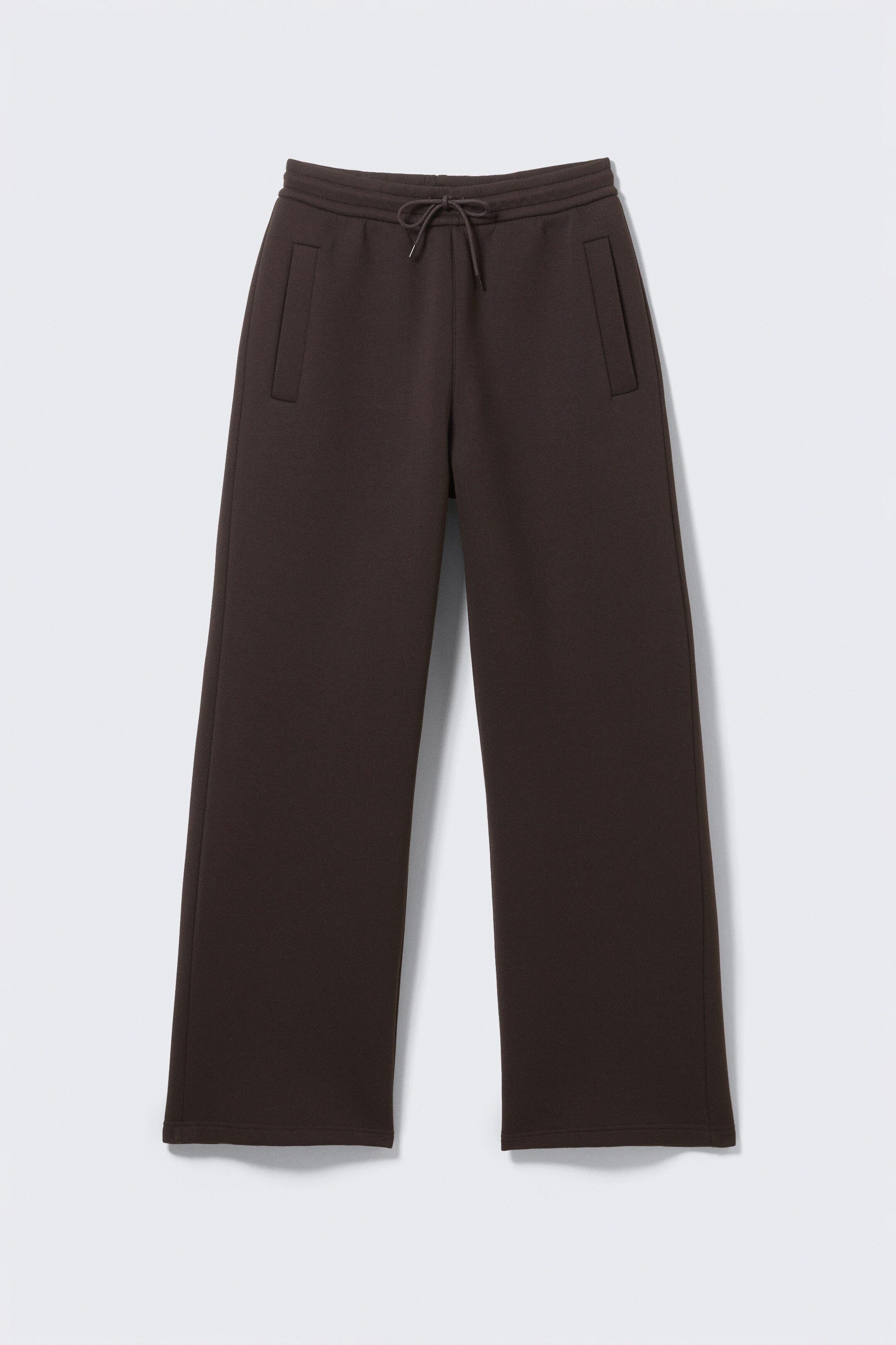 Agrandir l'image: Vue de FACE d'un pantalon de jogging ample de couleur marron foncé, confectionné en tissu scuba épais, avec une taille élastiquée munie d'un cordon de serrage noué, deux poches passepoilées à l'avant, et une silhouette décontractée.