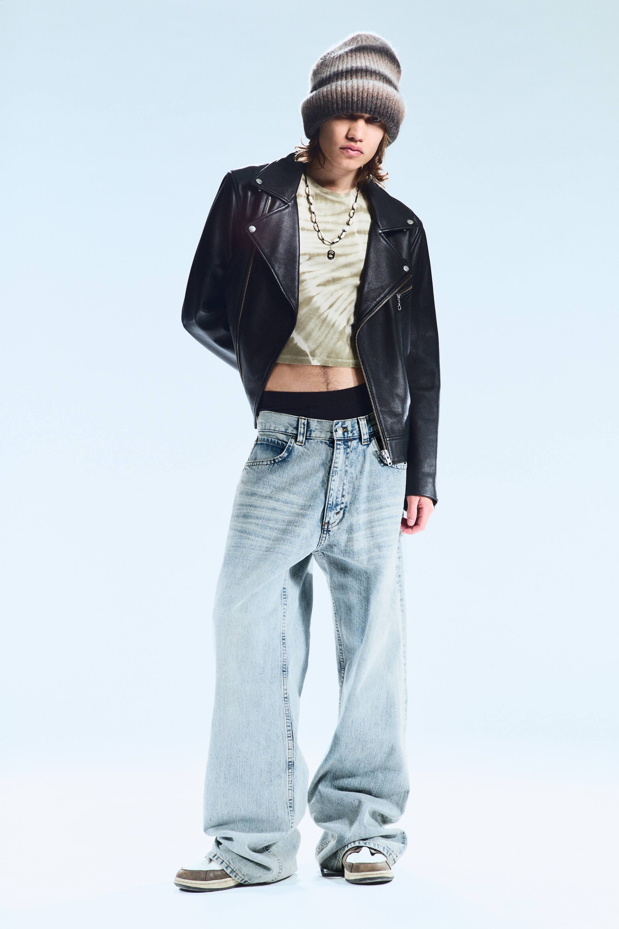 Größeres Bild anzeigen: Ein Outfit wird präsentiert, das eine gestreifte Strickmütze in Braun- und Grautönen, eine schwarze Leder-Bikerjacke und ein hellgrünes Tie-Dye-Crop-Top umfasst. Es werden hell gewaschene, weit geschnittene Baggy-Jeans getragen, die über braun-weißen Sneakers fallen, und eine silberne Kettenhalskette ist ebenfalls sichtbar.