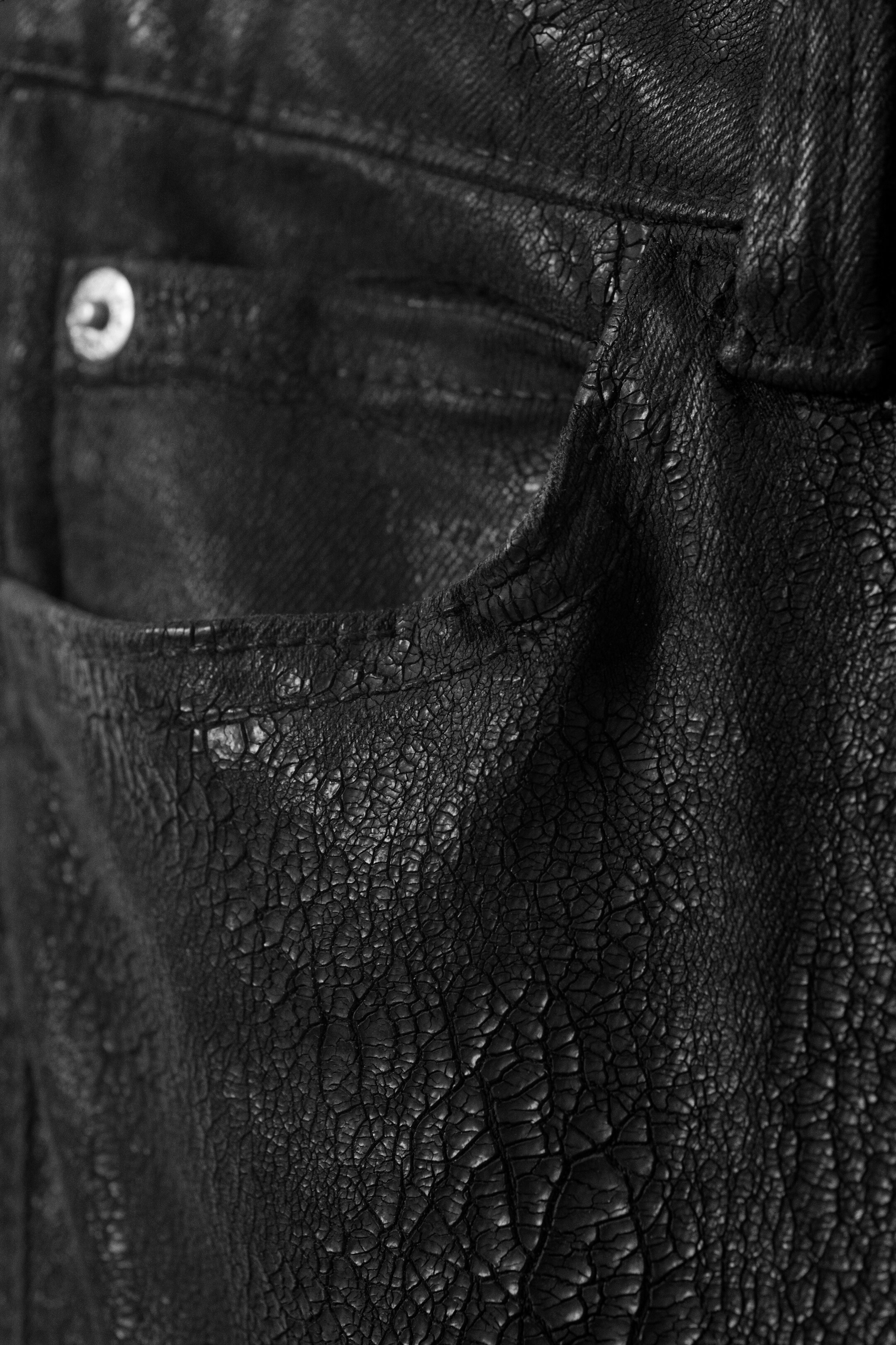 Agrandir l'image: Un pantalon noir présente une surface fortement craquelée et texturée sur le devant, avec une poche avant visible et une plus petite poche à monnaie. Un rivet argenté détaille le coin de la poche à monnaie.