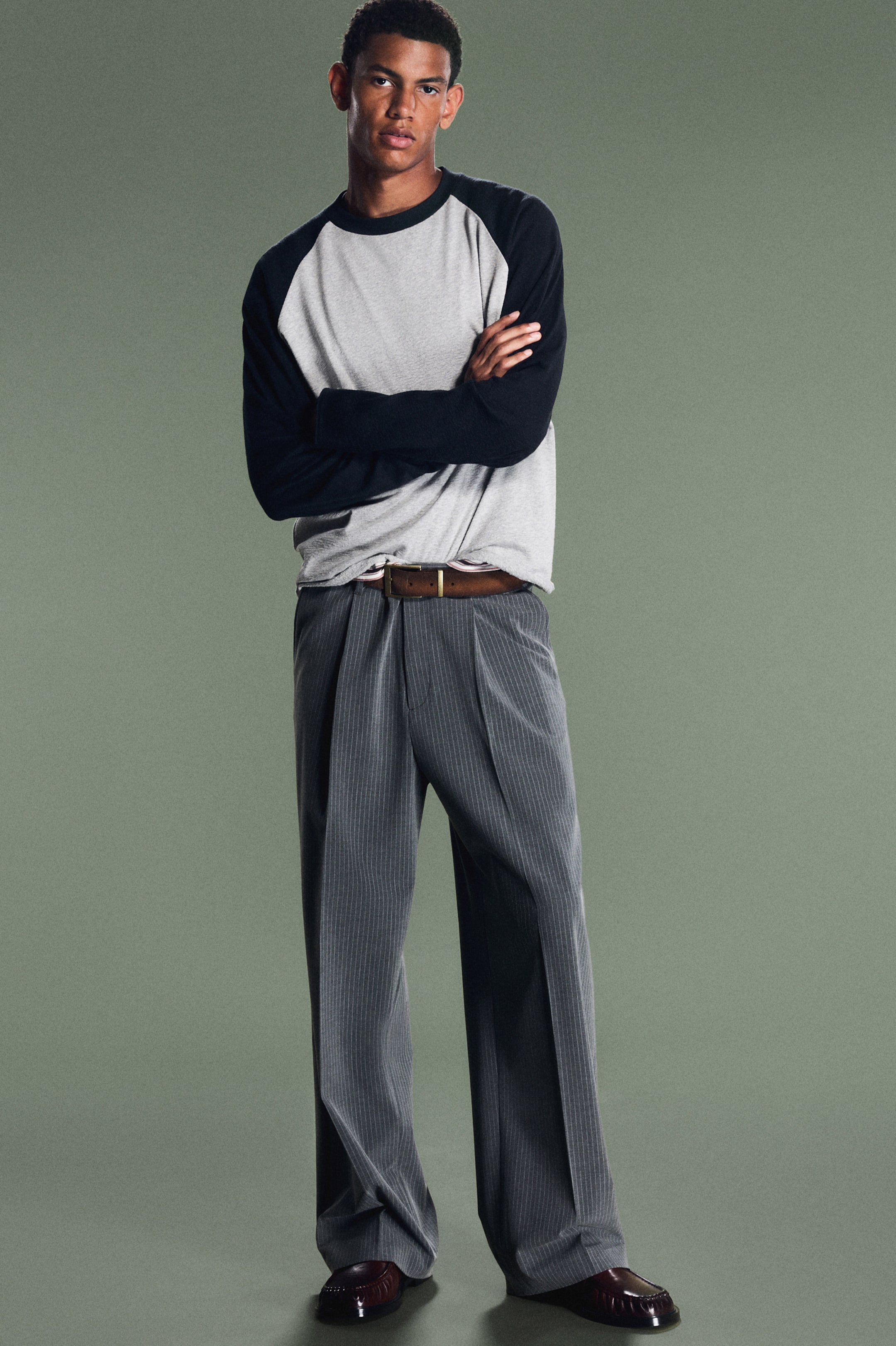 Vezi imaginea mai mare: Pantaloni supradimensionați, croiți pe corp, într-un model cu dungi fine gri, sunt purtați de model, având pliuri în față și dungi călcate la dungă. Pantalonii sunt asortați cu o curea din piele maro, un tricou raglan cu mânecă lungă gri și negru și loafers închiși la culoare.