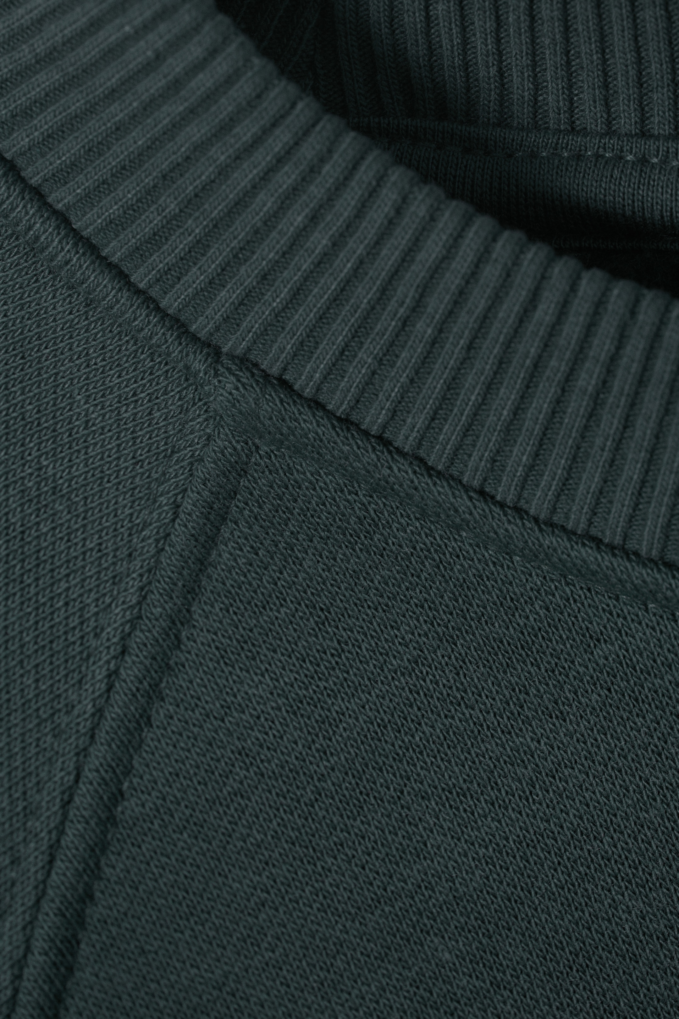 Se større billede: En mørkegrøn sweater præsenteres i en nærbillede, der fremhæver dens ribbede rundhals og den teksturerede strik i stoffet, med en synlig raglan-søm, der forbinder sektionerne.