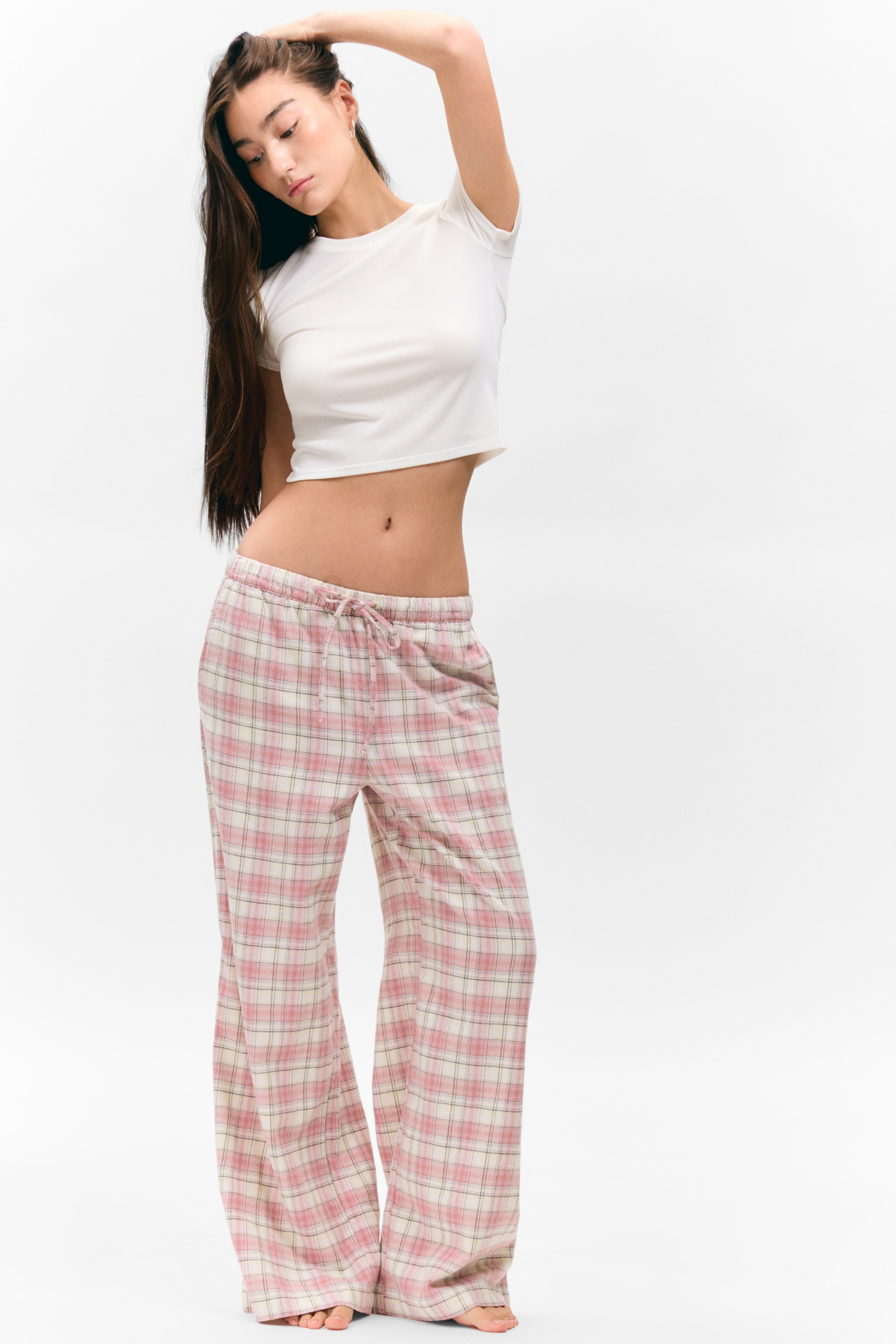 Größeres Bild ansehen: Eine Hose mit Karomuster zeigt ein pinkes, cremefarbenes und dünnes, dunkel umrandetes Muster und präsentiert eine lockere, weit geschnittene Passform aus einem frontalen Blickwinkel. Die Hose verfügt über einen elastischen Bund mit einer Kordelzugbindung vorne und wird mit einem weißen, kurzärmeligen Crop-Top getragen.