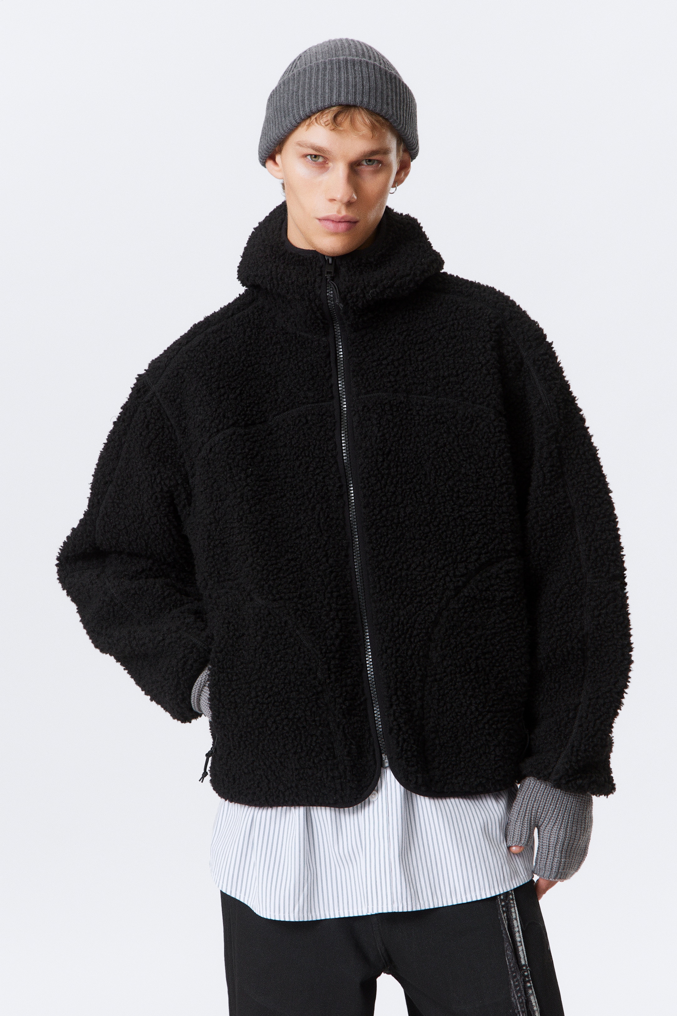 Visualizza immagine più grande: Una giacca in pile sherpa nero è rivolta in avanti, con una chiusura frontale con zip completa, un collo alto e due tasche laterali inserite con aperture curve. È indossata sopra una camicia a righe, con guanti grigi senza dita visibili ai polsi.