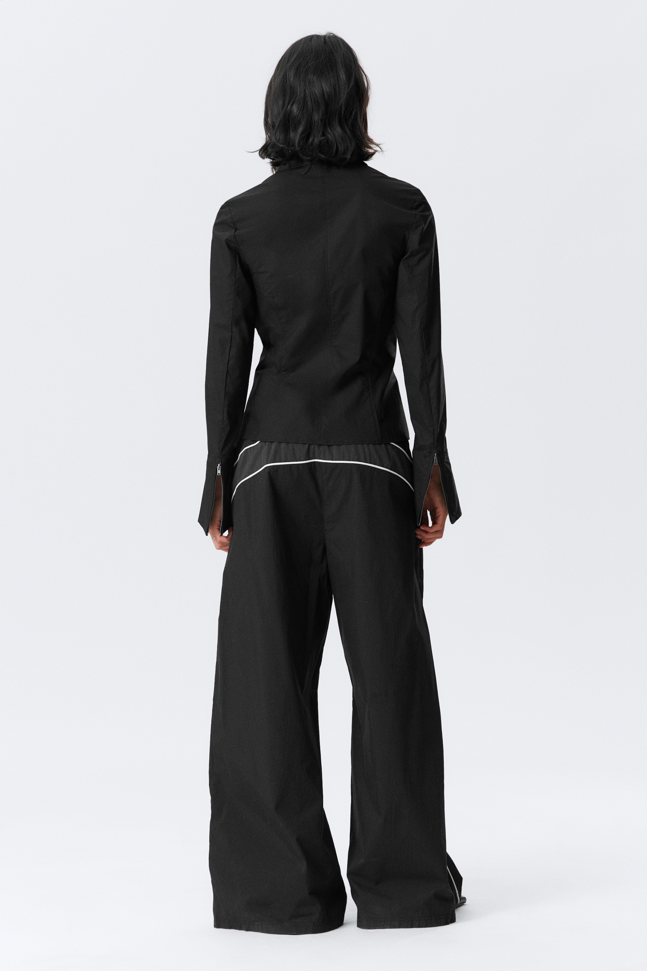 Größeres Bild anzeigen: Ein schwarzes zweiteiliges Outfit wird von hinten präsentiert, mit einem eng anliegenden Oberteil mit langen Ärmeln und Reißverschlussdetails an den Manschetten. Die dazu passenden Hosen mit weitem Bein zeigen einen horizontalen weißen Streifen um den Hüftbereich und einen vertikalen weißen Streifen entlang der Beinseite.