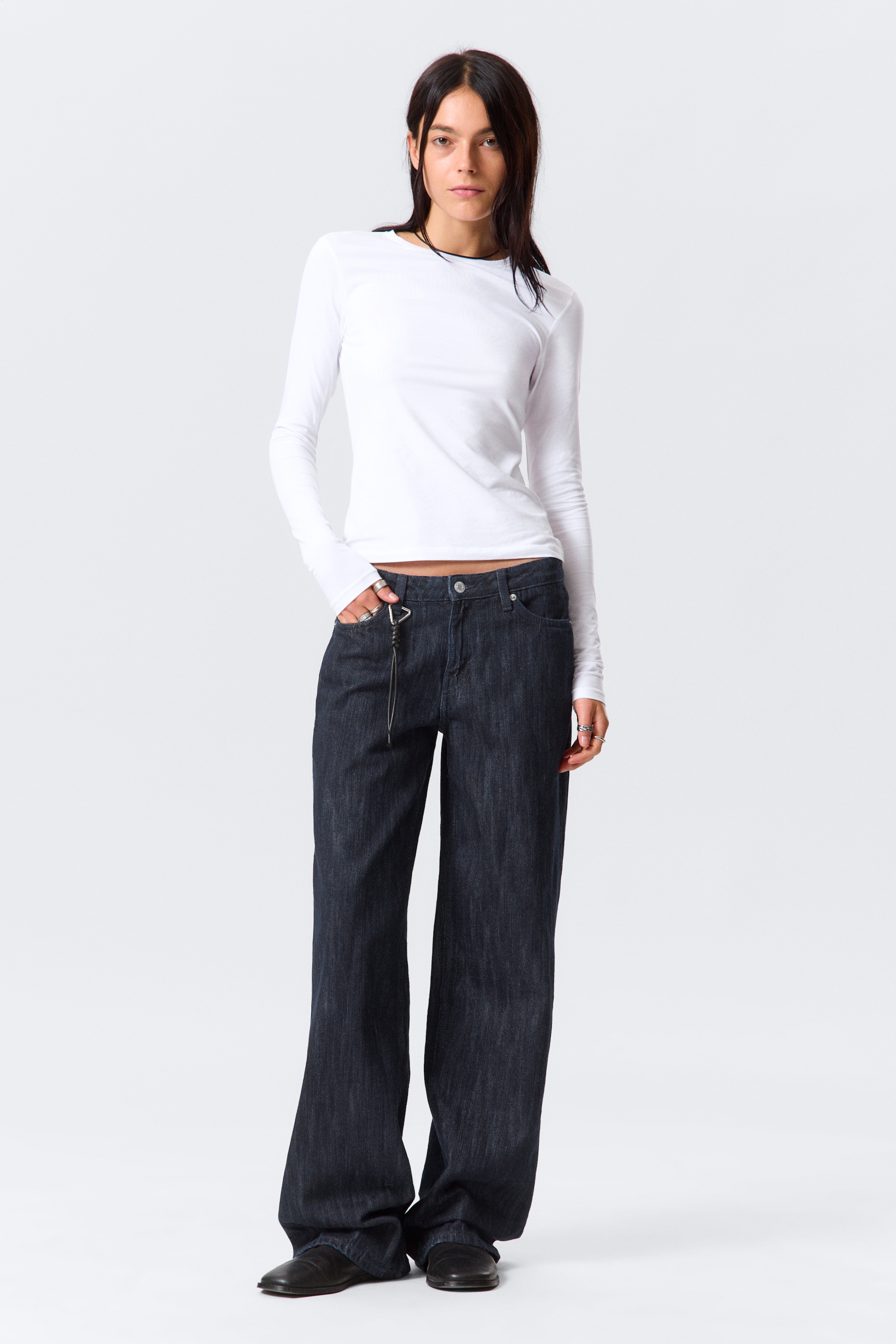 Grotere afbeelding bekijken: Een paar donker gewassen, wijde jeans wordt gepresenteerd, met een klassiek vijfzakkenontwerp en een kleine kettingaccessoire die aan een voorste riemlus is bevestigd. Een aansluitende witte top met lange mouwen, een ronde hals en verlengde mouwen die de handen bedekken, wordt gedragen met de jeans.