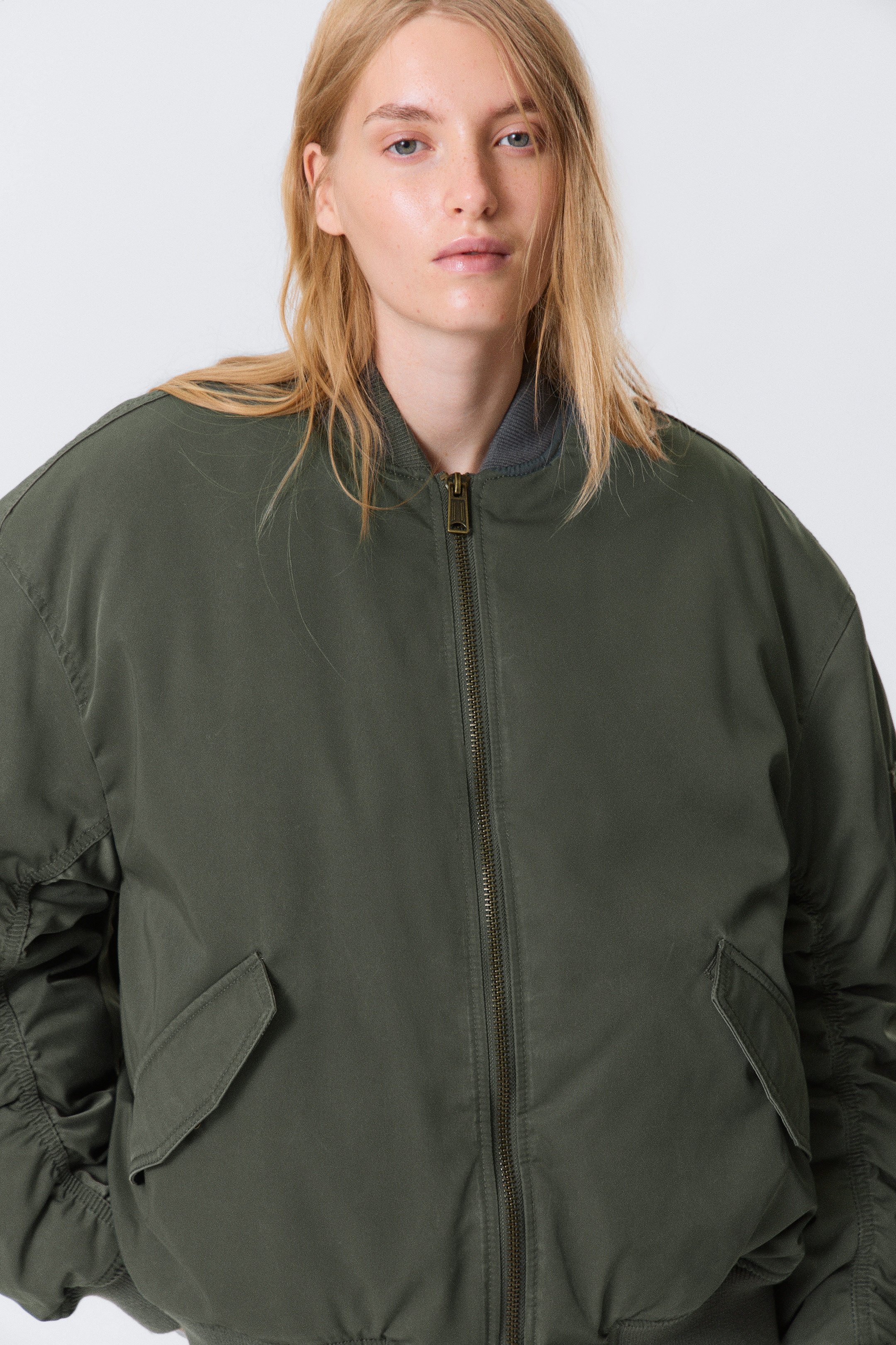 Visualizza immagine più grande: Una giacca bomber verde oliva viene presentata da un'angolazione frontale, mettendo in mostra la sua vestibilità rilassata. Presenta una chiusura con cerniera metallica a tutta lunghezza, un colletto a coste grigio, due tasche frontali con patta e dettagli arricciati sulle maniche.