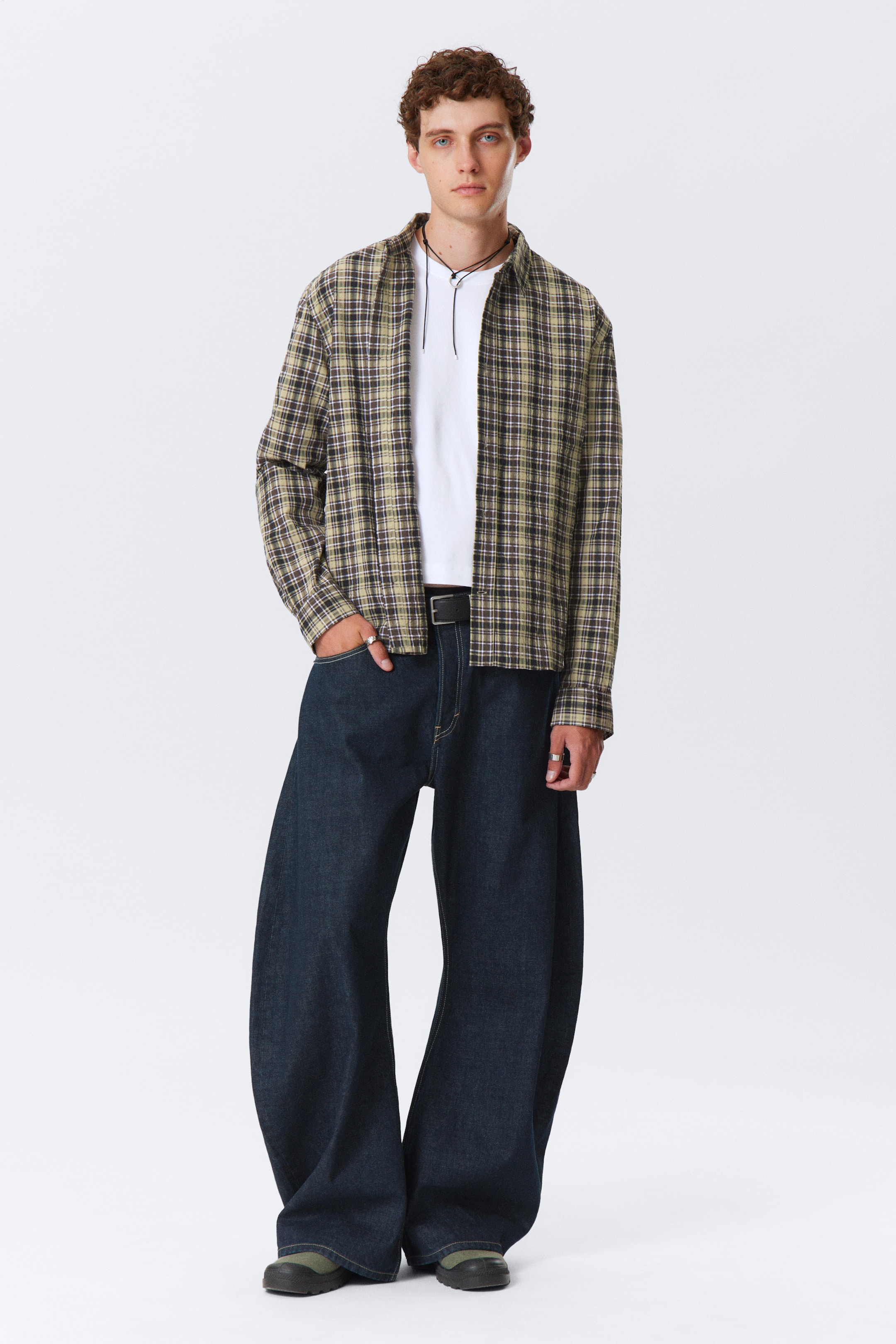 Visa större bild: En modell presenterar en avslappnad outfit, med en löst sittande, uppknäppt rutig skjortjacka i bruna, gröna och svarta toner, lager på lager över en vit t-shirt. Outfiten kompletteras med mörkblå, extremt vida jeans, som är åtdragna med ett svart bälte, och mörkgröna slip-on skor.