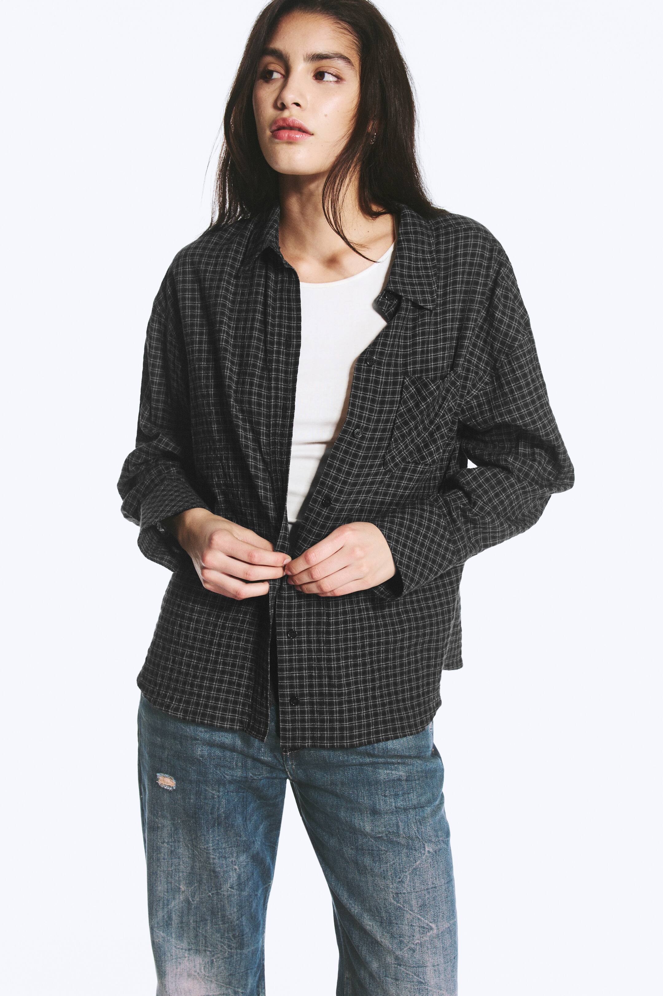Se større billede: En oversized mørkternet skjorte med klassisk krave og en brystlomme bæres af modellen over en hvid T-shirt og blå jeans. Modellen holder og justerer skjortens frontlukning med knapper på den afslappede pasform.