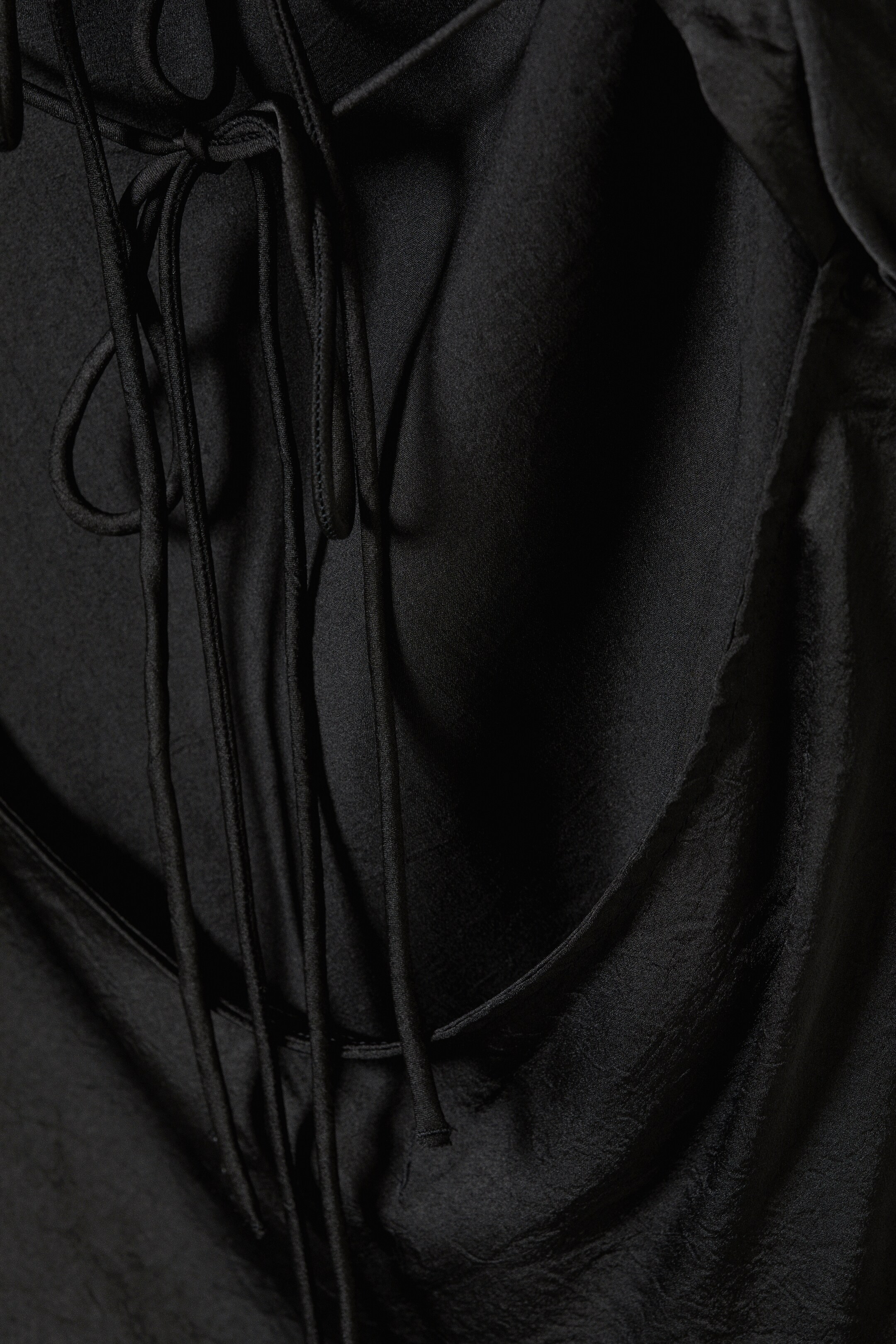 View larger image: A black garment is showing its back, featuring multiple thin cords gathered and tied at the top, creating a draped effect in the lustrous fabric below.