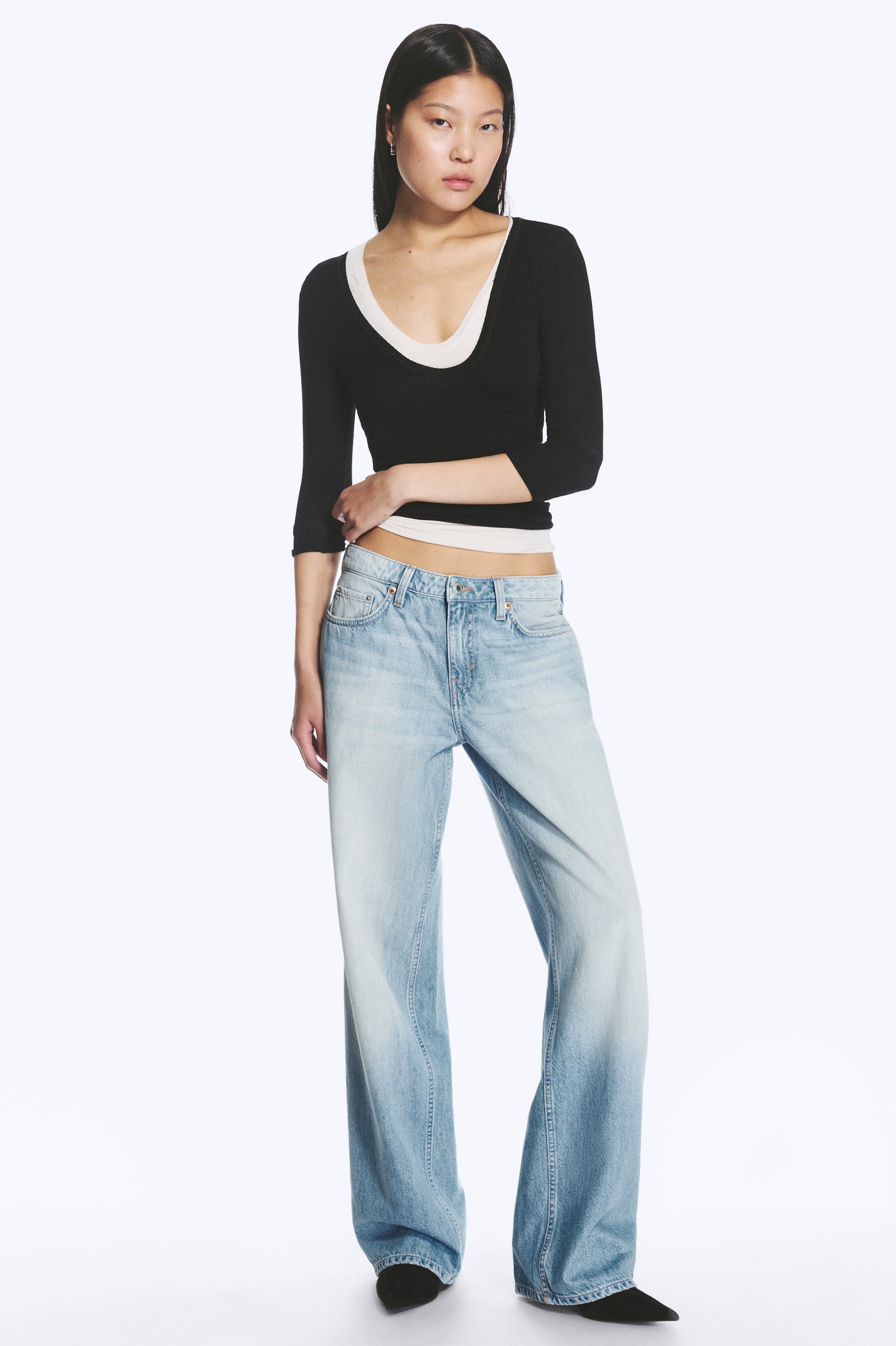 Se større billede: Jeans med lav talje, løs pasform og brede ben i lysvasket, stiv denim bæres af modellen. Jeansene har en lang længde, der falder i folder omkring fødderne, og har synlige frontlommer. Modellen rører let ved jeansenes linning med den ene hånd.