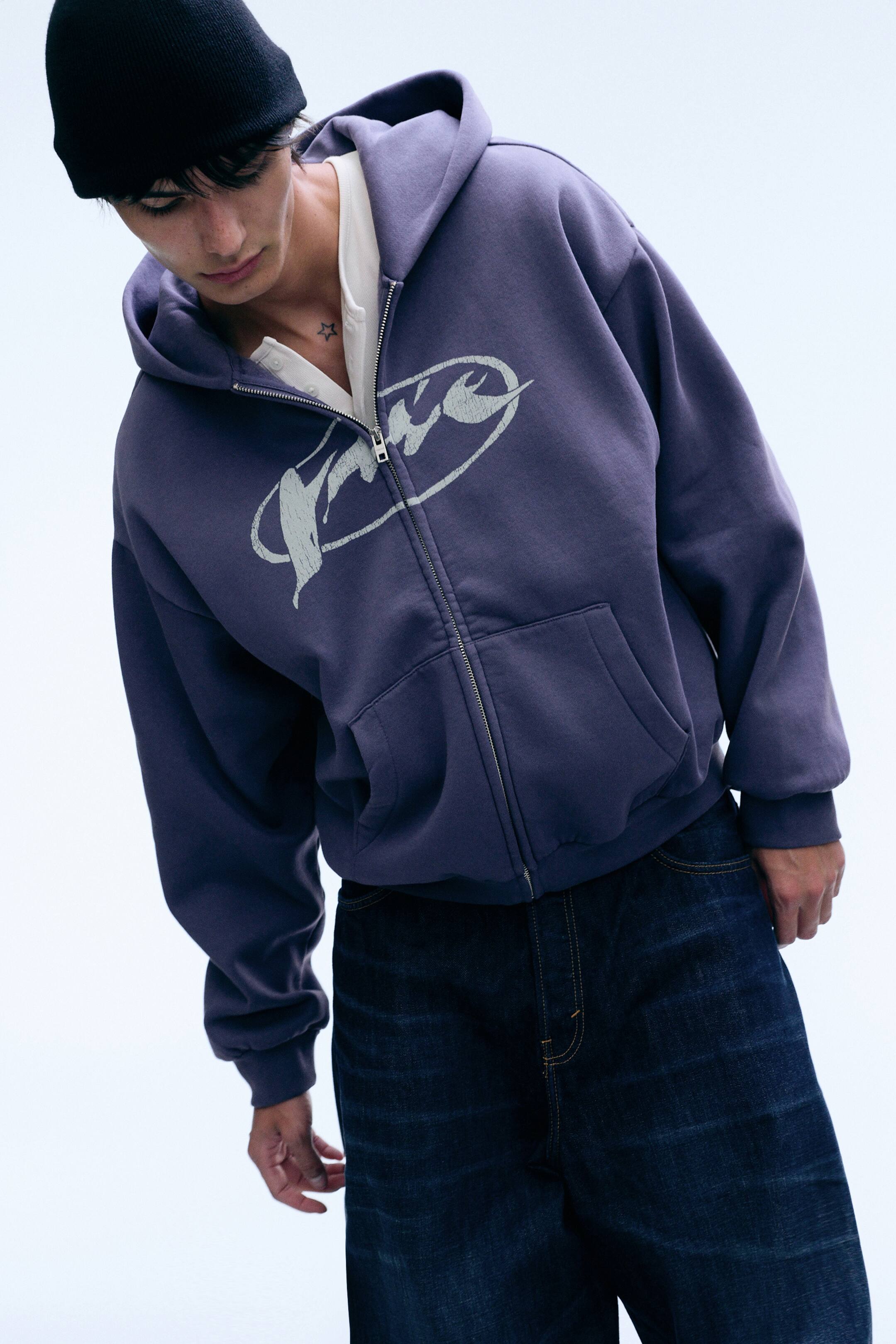 Vis større bilde: En lilla, løstsittende zip-hoodie med grafisk trykk bæres av modellen over en offwhite henley-skjorte og mørke jeans. Hoodien har et falmet grafisk trykk på brystet, en delvis glidelåsåpning foran, droppede skuldre og en delt kengurulomme.