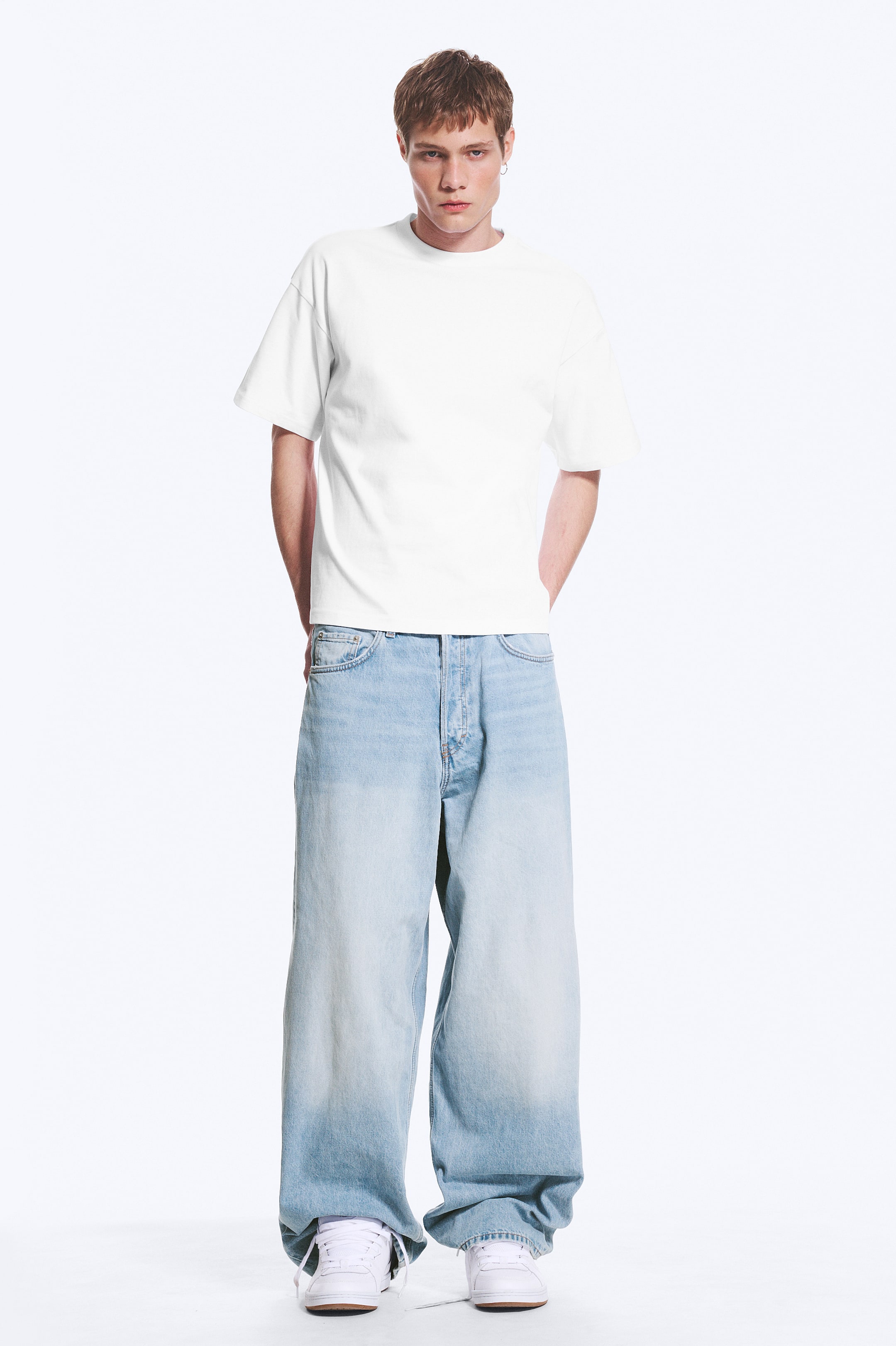 Visualizza immagine più grande: La modella indossa una T-shirt bianca oversize con scollo rotondo, spalle cadenti e maniche ampie. La T-shirt è infilata in un paio di jeans oversize azzurri, e la modella è in piedi con le mani nelle tasche.