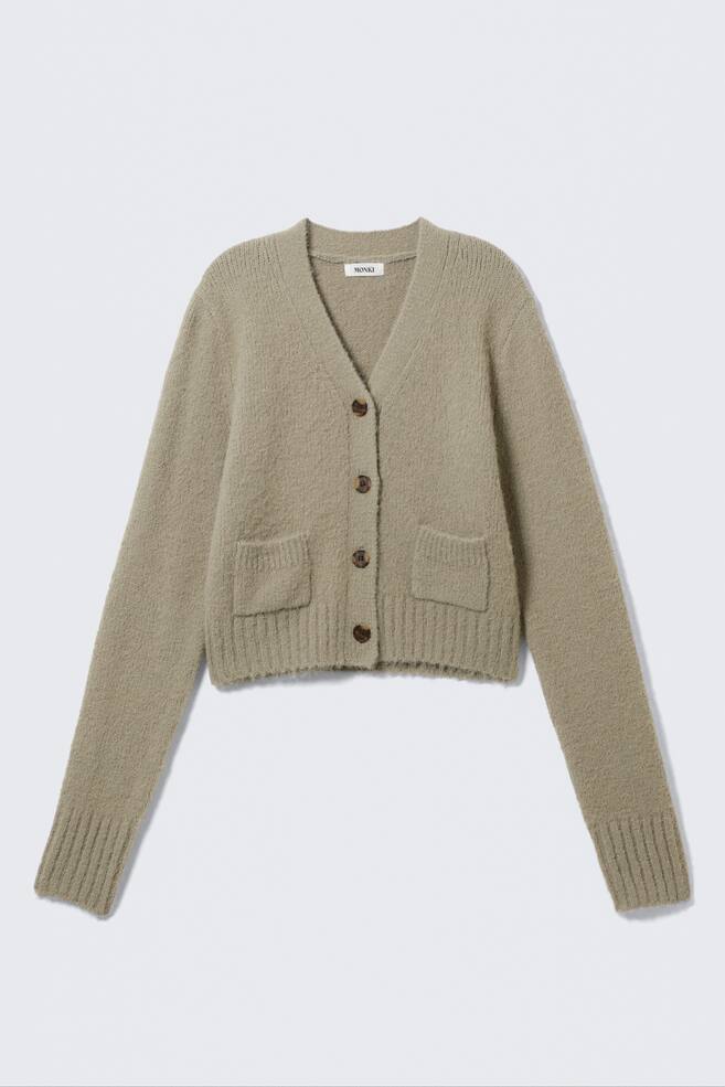 Un cardigan lavorato a maglia verde salvia e peloso è presentato frontalmente, con scollo a V, quattro bottoni marroni e due tasche applicate. È progettato con polsini a coste e un orlo a coste, in uno stile corto. - 1