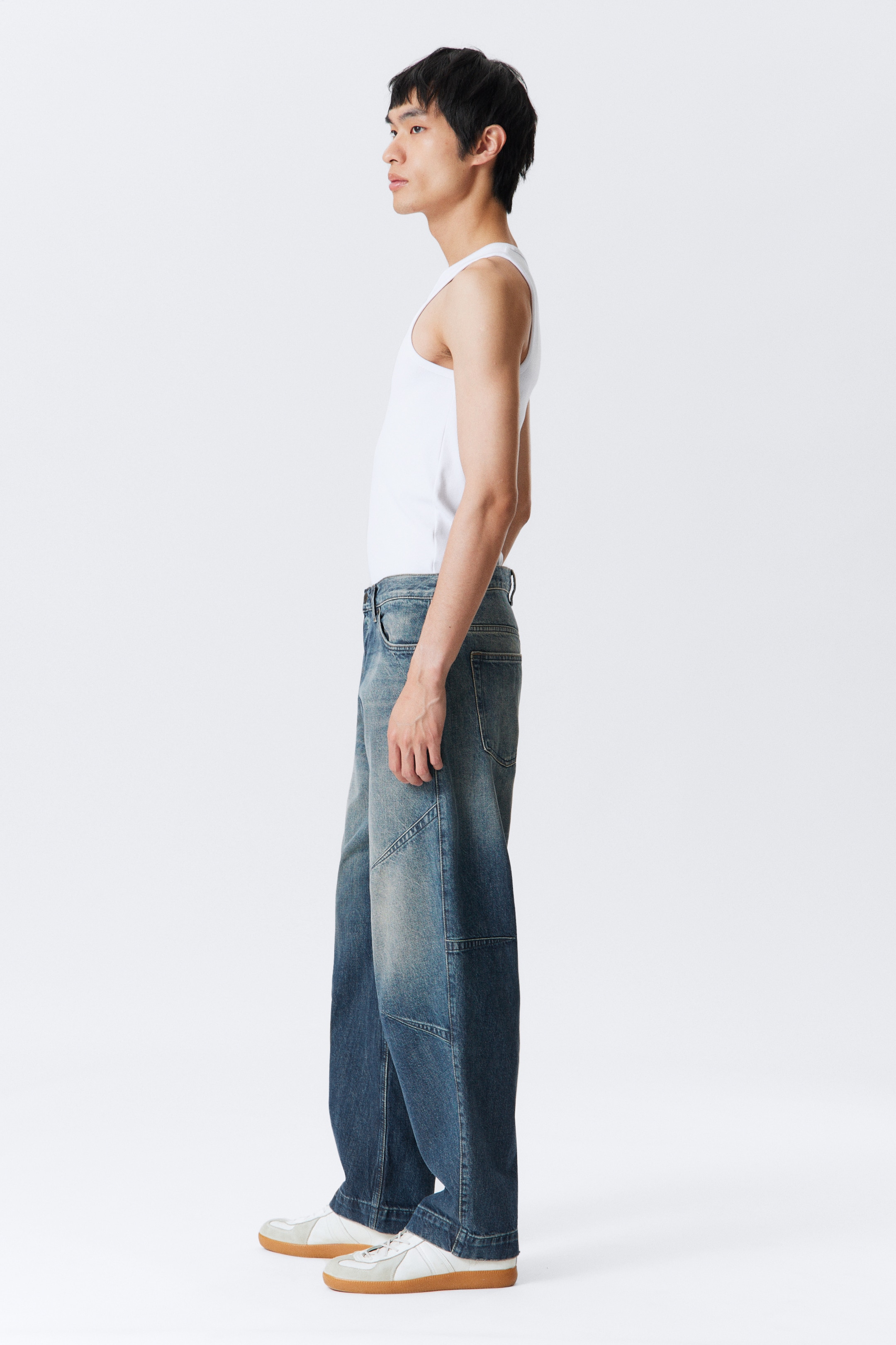 Agrandir l'image: Un jean bleu à coupe ample est porté par le mannequin, avec une taille basse et une jambe qui semble fuselée. Des détails de pinces en biais sculptent la silhouette de la jambe, et le denim présente des tons de bleu variés avec une apparence rigide.