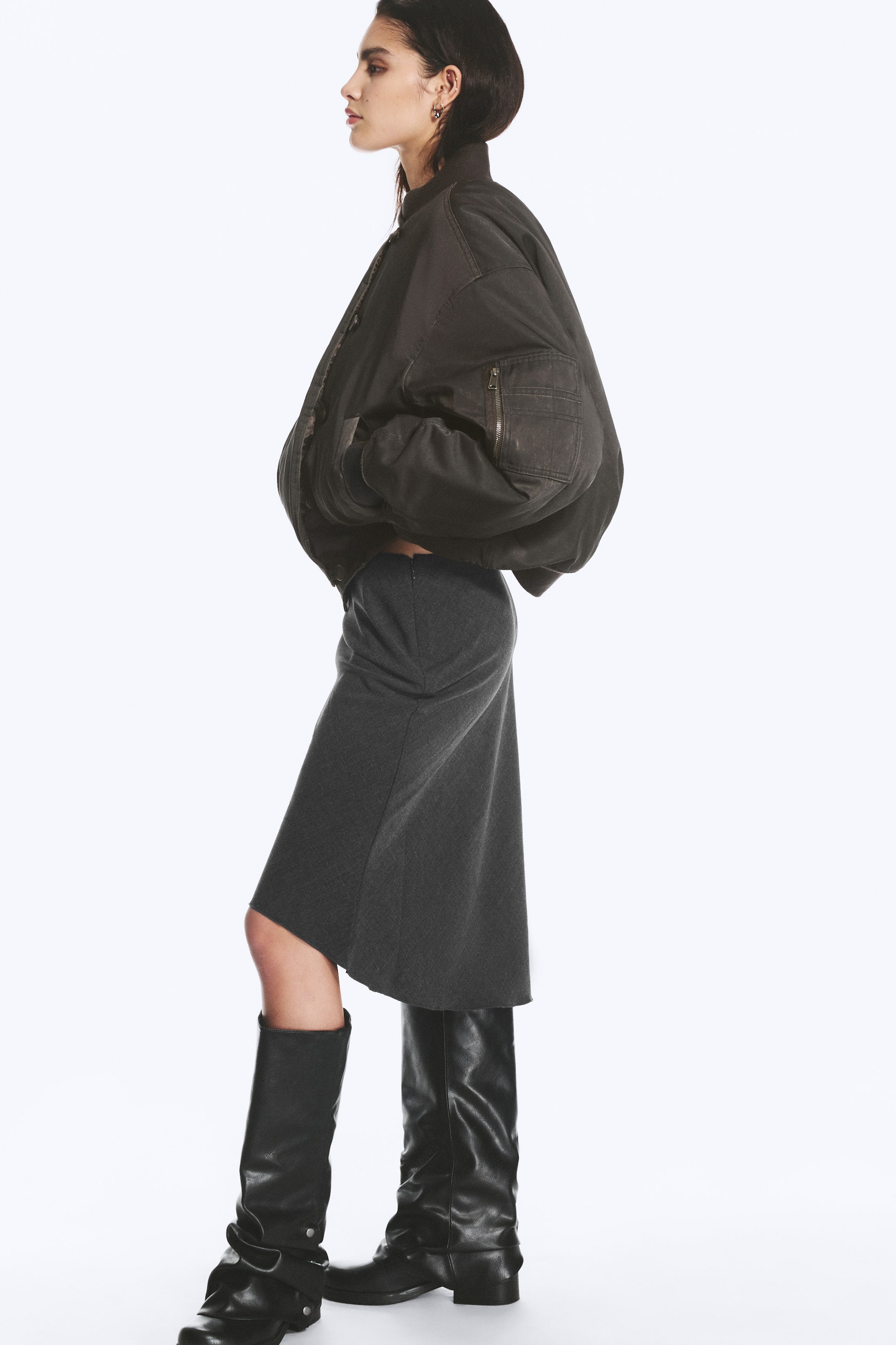 Agrandir l'image: Une veste bomber oversize en tissu charbon délavé est portée par le mannequin, qui se tient de profil, les mains glissées dans les poches latérales. La veste présente une silhouette volumineuse, des finitions côtelées et une poche zippée sur la manche, et elle est associée à une jupe midi gris foncé ainsi qu’à de hautes bottes noires en cuir.