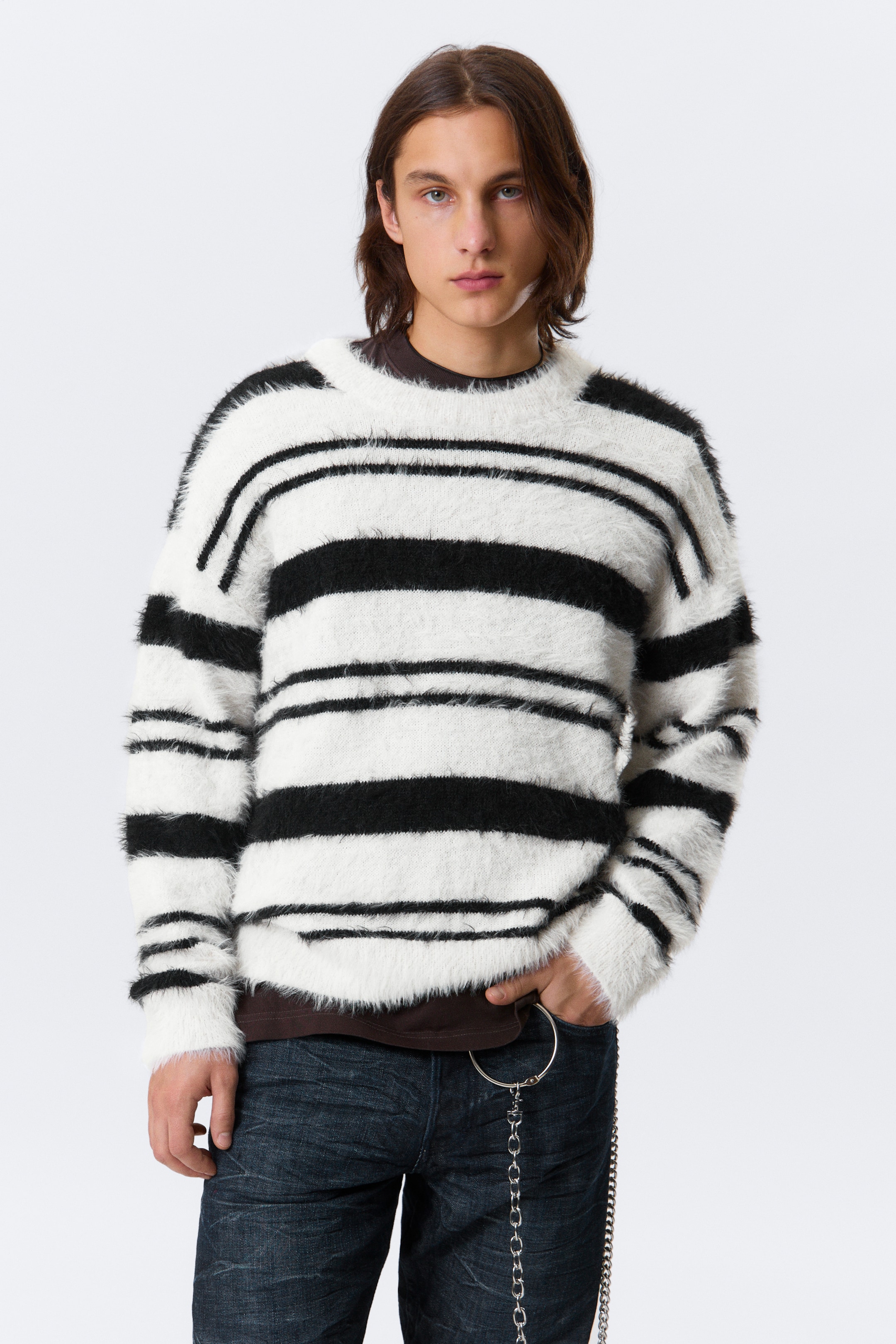 Größeres Bild anzeigen: Ein flauschiger Strickpullover wird präsentiert, der schwarze und weiße horizontale Streifen in unterschiedlichen Breiten aufweist. Der Pullover zeigt einen Rundhalsausschnitt und eine lockere Passform und wird über einem sichtbaren dunkelbraunen T-Shirt getragen.
