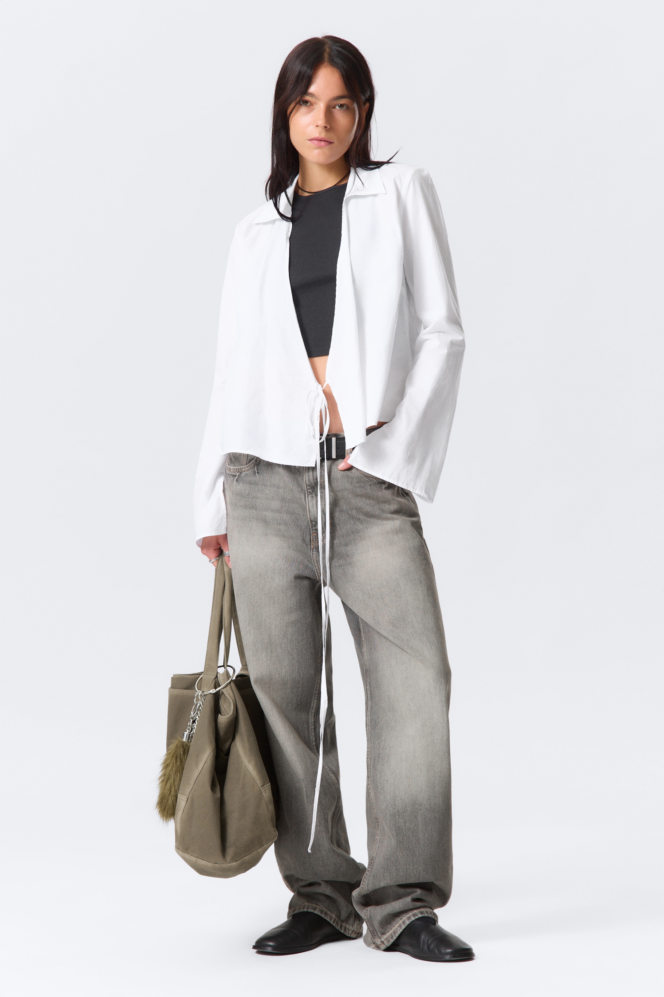 Visa större bild: Outfiten, presenterad framifrån, har en vit kortärmad skjorta med krage som bärs öppen och avslöjar en svart kort topp under. Skjortan har långa band som hänger från framsidan och är stylad med blekta gråa vida jeans, åtdragna i midjan med ett svart bälte. Svarta platta skor kompletterar looken, medan en olivgrön tote-väska med en pälscharm bärs.