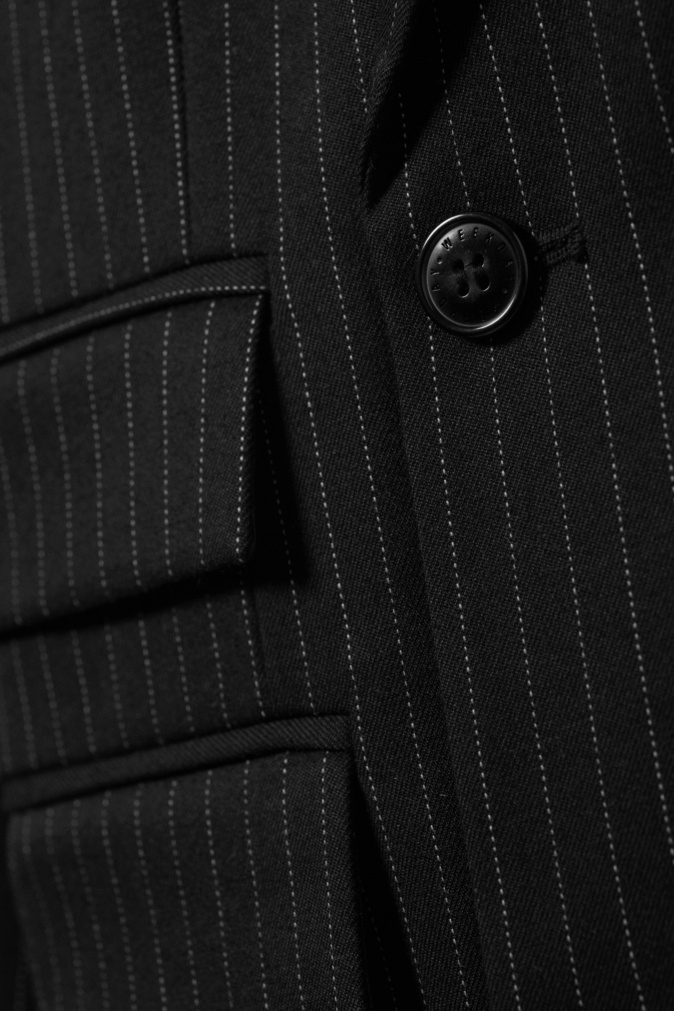 Agrandir l'image: Un blazer noir à rayures est présenté, affichant son devant avec deux poches à rabat rectangulaires et un bouton noir proéminent. Le bouton présente un texte en relief.