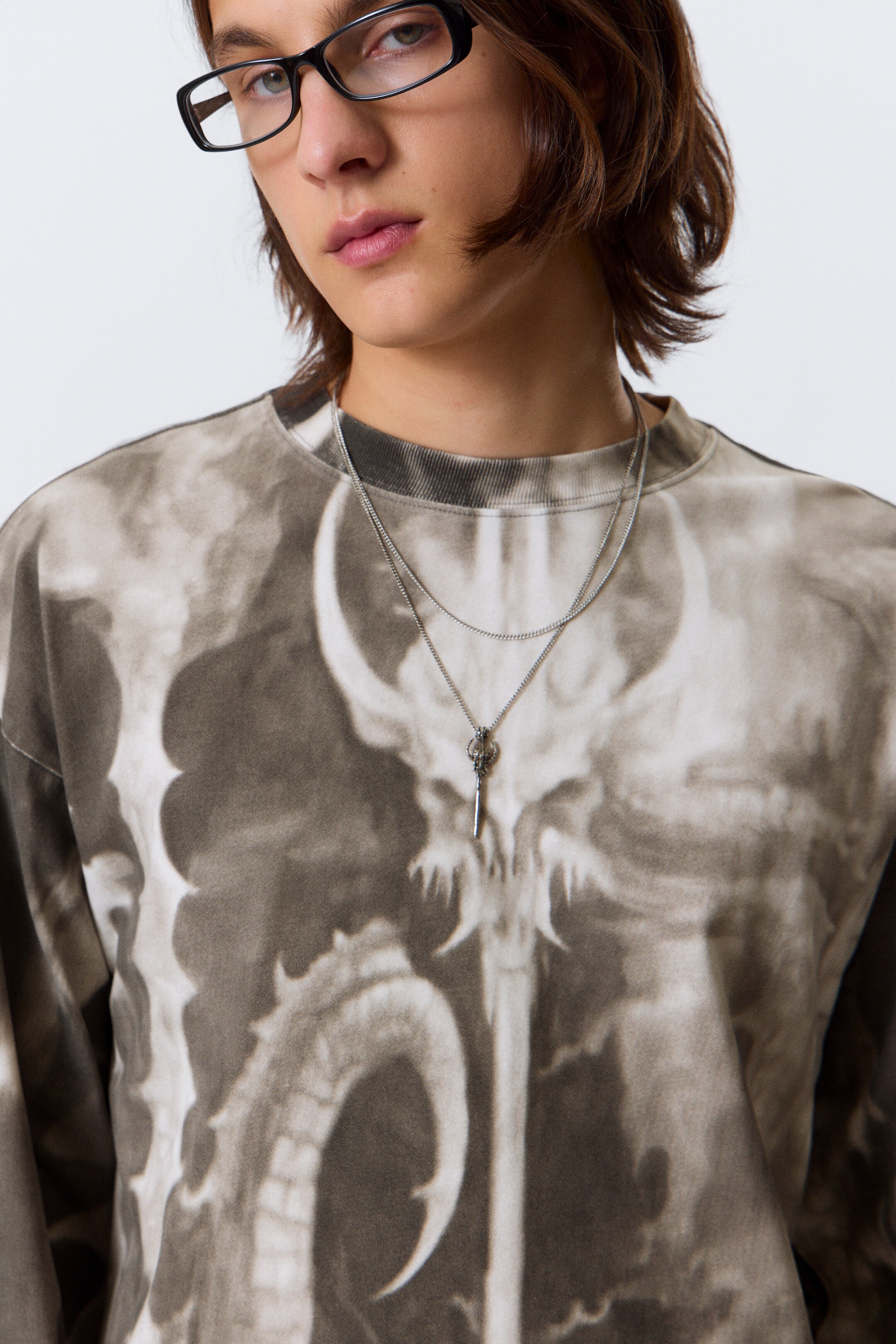 Agrandir l'image: Un t-shirt à manches longues et col rond affiche un motif tie-dye noir et marron délavé avec un graphique squelettique central. Un collier à deux couches de ton argenté repose sur le t-shirt, avec une chaîne à maillons et un pendentif en forme de clé.