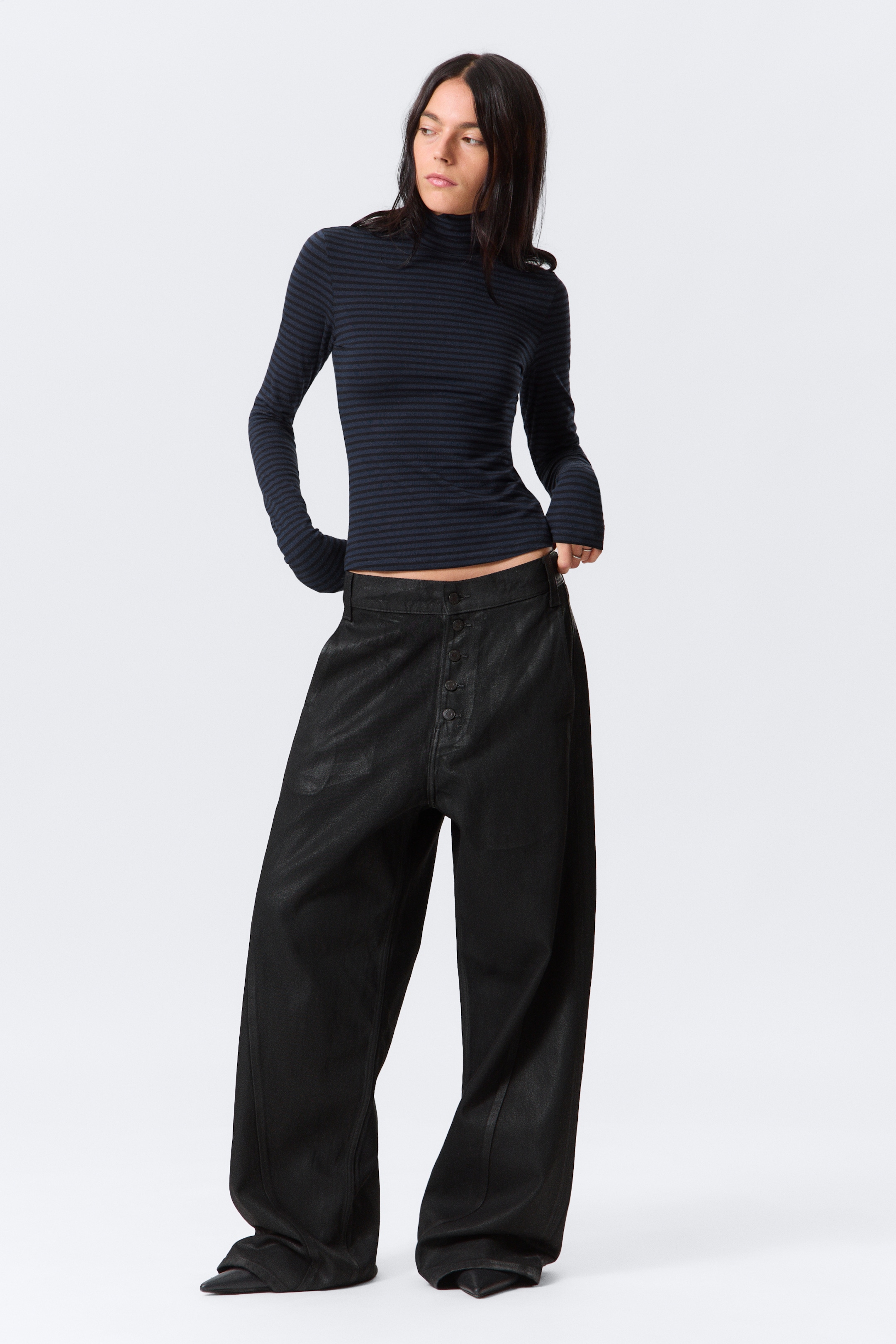 Grotere afbeelding bekijken: Een aansluitende coltrui met een algeheel gestreept patroon en een dubbelgelaagde kraag wordt gedragen door het model. Het model houdt de onderste zoom van de top met beide handen vast en trekt deze iets van het lichaam af.