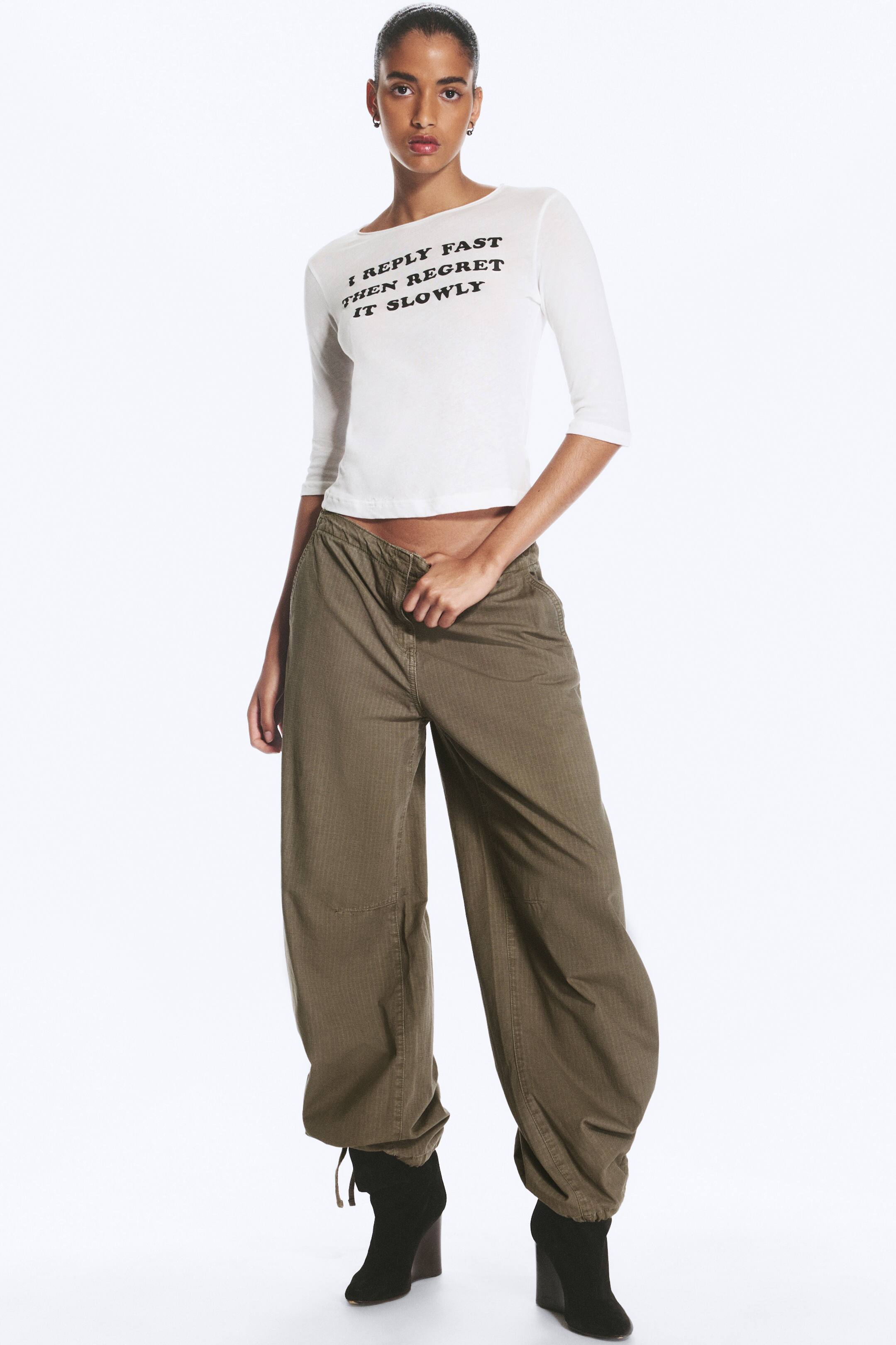 Agrandir l'image: Le mannequin porte un pantalon taille basse vert olive, avec une silhouette ample et baggy, et des ourlets resserrés par un cordon de serrage. Le mannequin ajuste la taille du pantalon, porté avec un top court blanc et des bottines noires à talons.