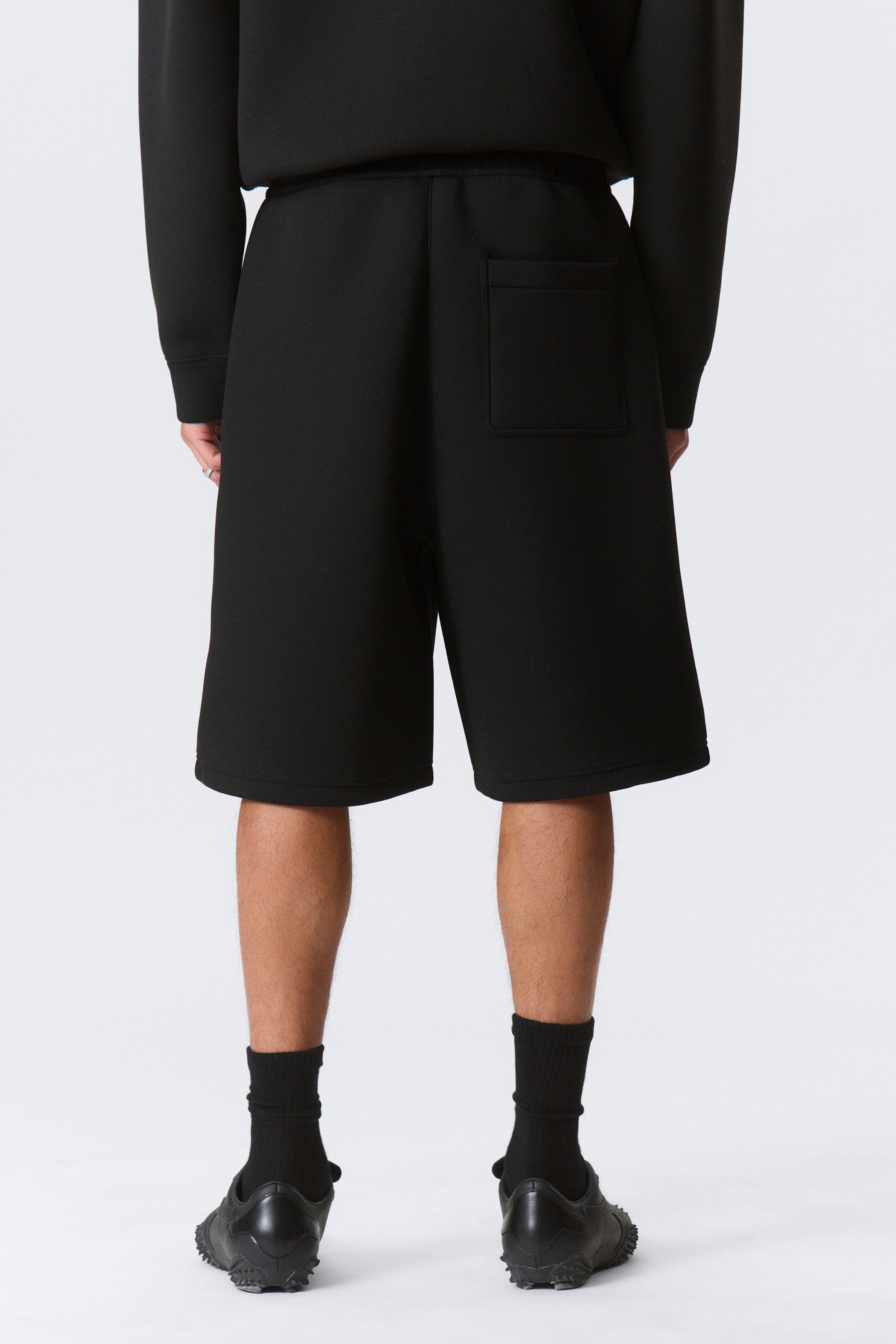 Vis større bilde: Svart shorts presenteres fra baksidevisning, med en elastisk linning og en enkelt lomme på høyre side. Shortsene faller over knærne med en enkel kant.