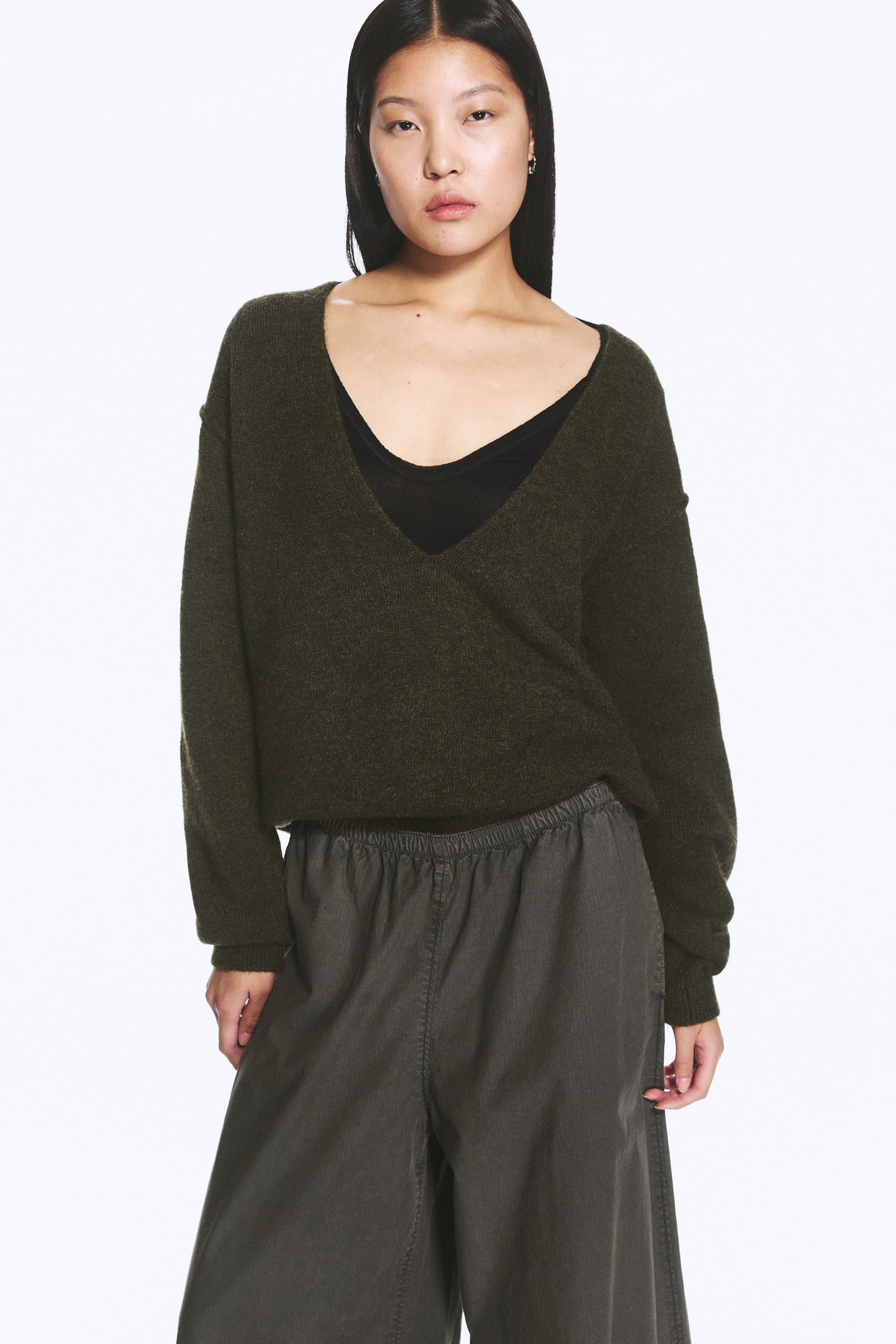 Agrandir l'image: Un pull vert foncé à col en V, à manches longues et à coupe décontractée, est porté par le mannequin, qui le superpose à un haut noir visible au niveau de l’encolure.