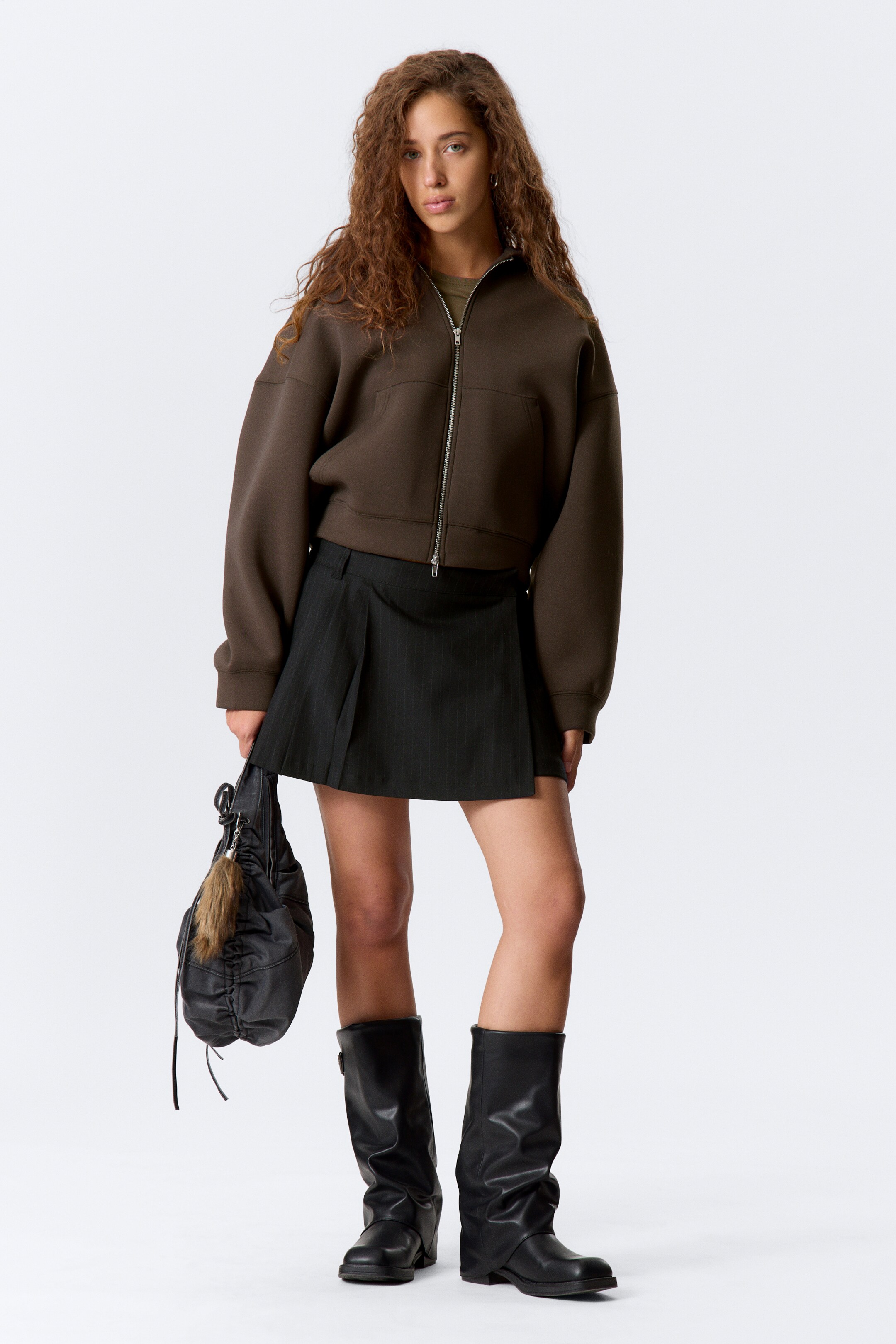 View larger image: A brown oversized zip-up jacket is featuring a standing collar, dropped shoulders, a full-length silver zipper, and two front welt pockets. It is paired with a black pinstripe pleated mini skirt, showcasing pleats and a side buckle detail, and black knee-high slouchy boots.