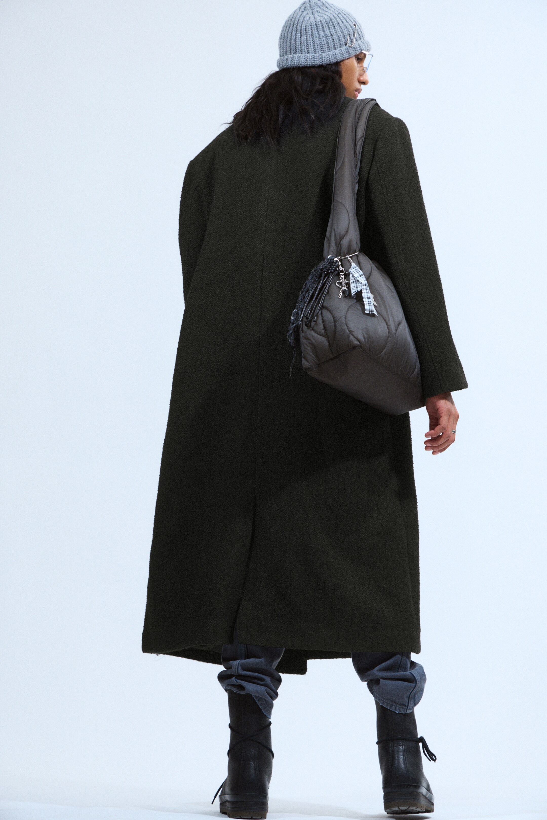Ingrandisci l'immagine: Un cappotto lungo di colore antracite scuro viene visto da dietro, caratterizzato da un tessuto strutturato e da una vestibilità ampia e oversize. Si estende oltre le ginocchia e mostra uno spacco visibile sul retro dell'orlo.