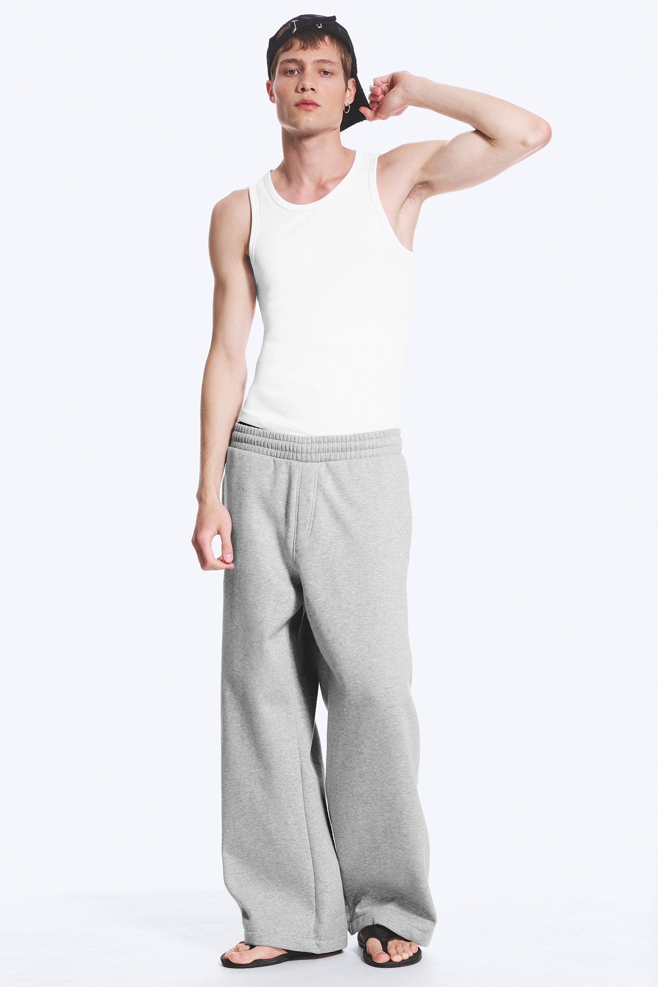 Vezi imaginea mai mare: Un maiou alb cu dungi este prezentat, având un guler rotund și un design mulat, băgat în pantaloni de trening gri. Pantalonii de trening au o talie elastică cu încrețituri, buzunare laterale vizibile și o croială largă și lejeră, care se adună la glezne.
