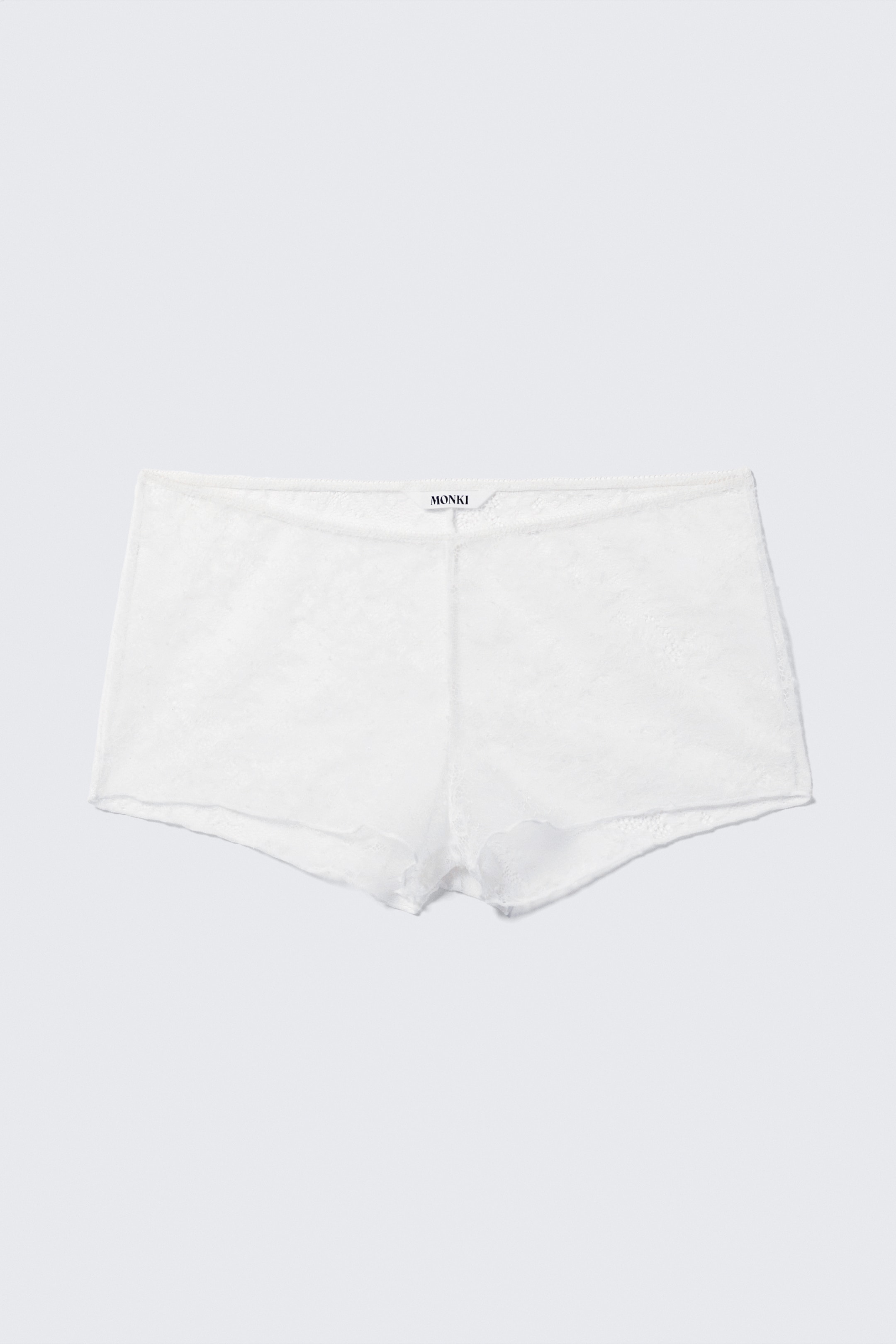 Größeres Bild anzeigen: Weiße Boyshorts liegen flach, von vorne präsentiert, und sind aus einem durchsichtigen Spitzenstoff gefertigt, der ein dezentes Blumenmuster zeigt. Ein 'MONKI'-Etikett ist in der Mitte des elastischen Bunds sichtbar, und sowohl der Bund als auch die Beinöffnungen sind mit zarten, gewellten Kanten versehen.