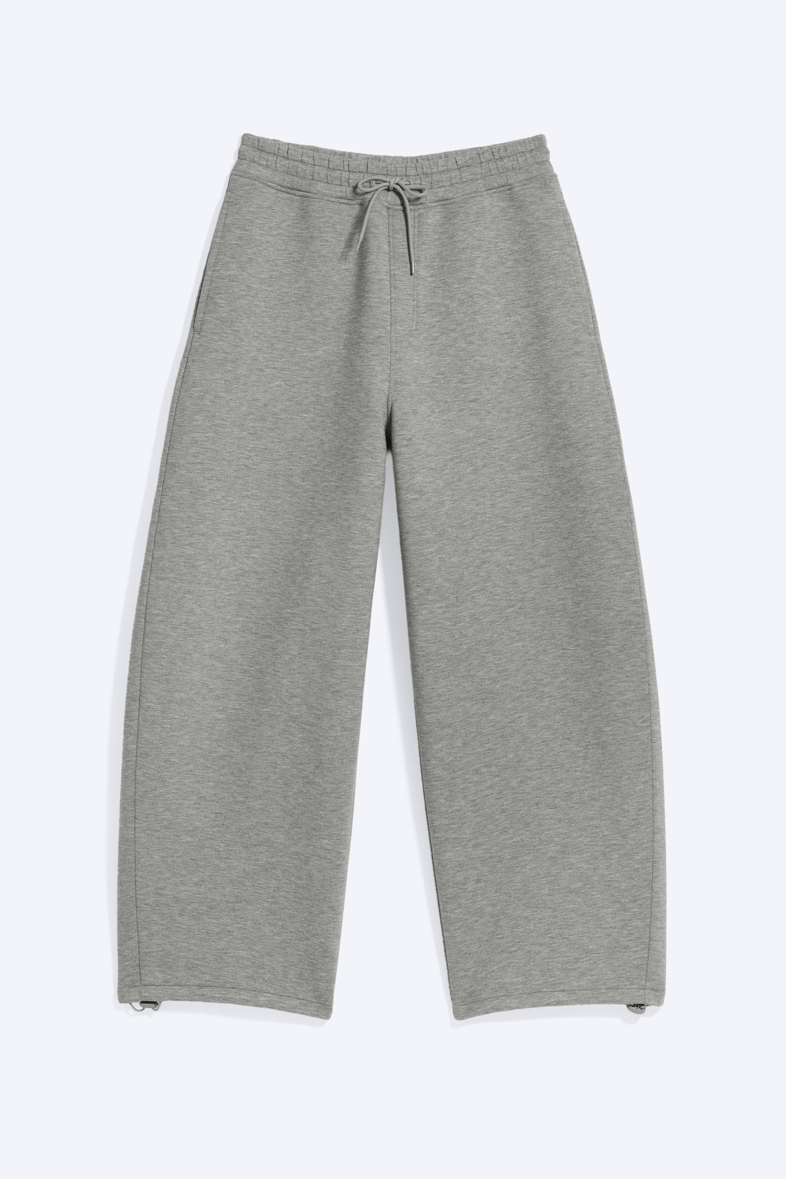 Pantalonii de trening gri cu cracul larg sunt așezați plat, având o talie elastică cu șnur legat și buzunare laterale în cusătură. Pe interiorul tivurilor drepte se observă mici cleme. - 1