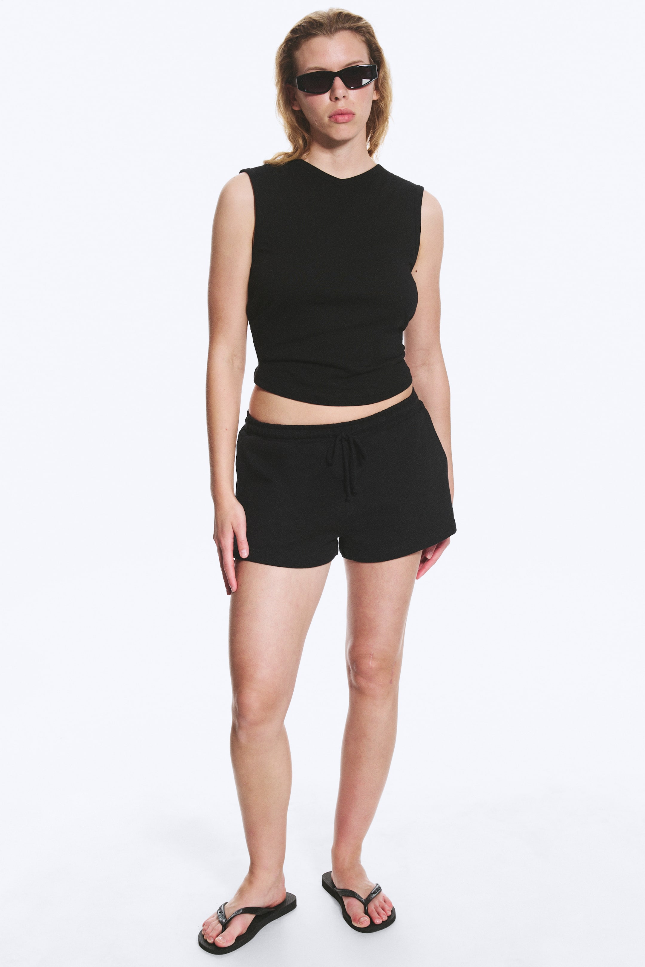 Agrandir l'image: Un haut noir sans manches et un short noir sont présentés de face. Le haut ajusté sans manches présente un col rond et un ourlet court, tandis que le short décontracté comprend une taille élastiquée avec un cordon de serrage visible et des poches latérales.