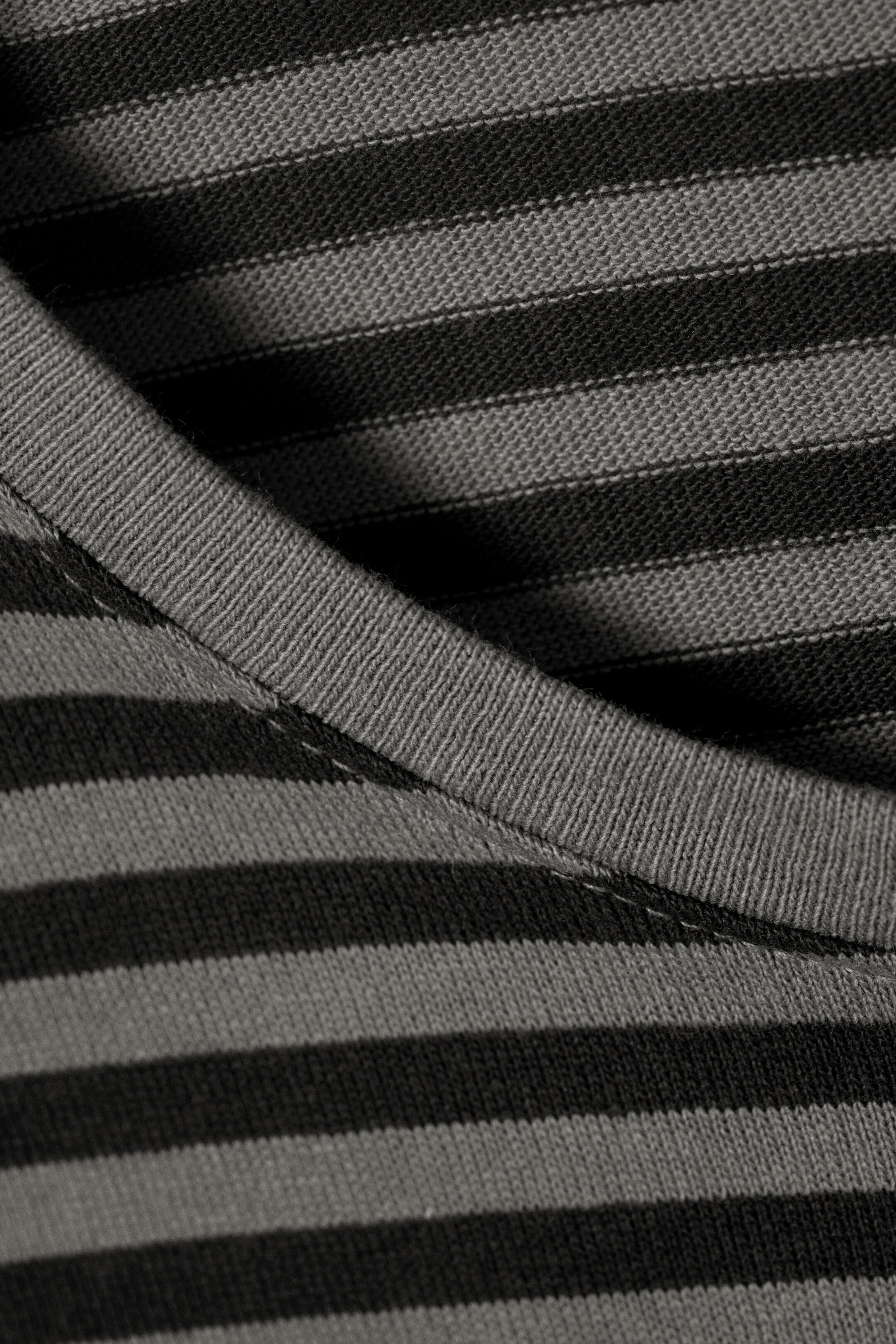 View larger image: A knit garment featuring horizontal dark gray and medium gray stripes is shown in close-up. A ribbed neckline or collar band is curving across the top right, displaying its texture and stitching.
