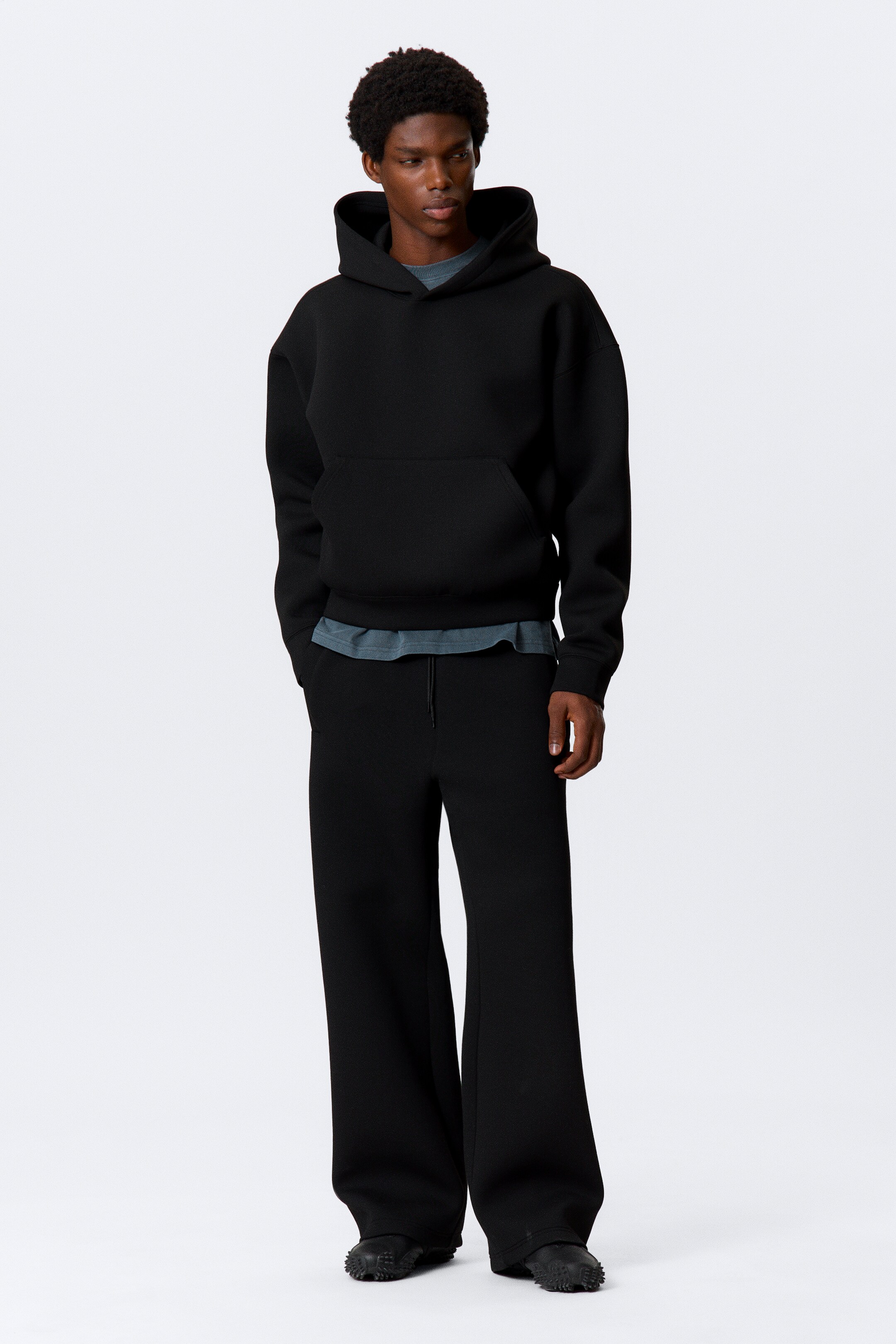 Grösseres Bild ansehen: Ein schwarzer oversized Kapuzenpullover ist nach vorne gerichtet, mit einer Kapuze, langen Ärmeln und einer vorderen Kängurutasche, darunter lugt ein graues T-Shirt hervor. Passende schwarze weite Hosen sind mit einem Kordelzugbund ausgestattet.