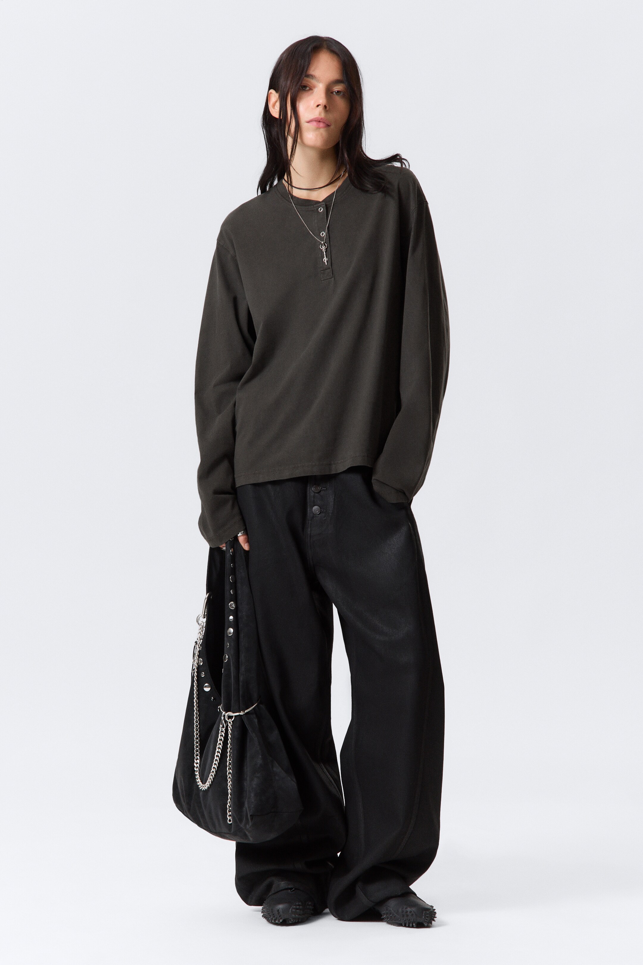 Grotere afbeelding bekijken: Een donkergrijs Henley-shirt met lange mouwen en een ontspannen, oversized pasvorm wordt gepresenteerd, met een drieknoopssluiting bij de halslijn en losse mouwen. Het wordt gedragen met loszittende zwarte wijde broek met een zichtbare knoopsluiting aan de voorkant en een subtiele glans, geaccessoriseerd met een zwarte slouchy tas versierd met zilveren studs en meerdere kettingen, en donkere, getextureerde platte schoenen.