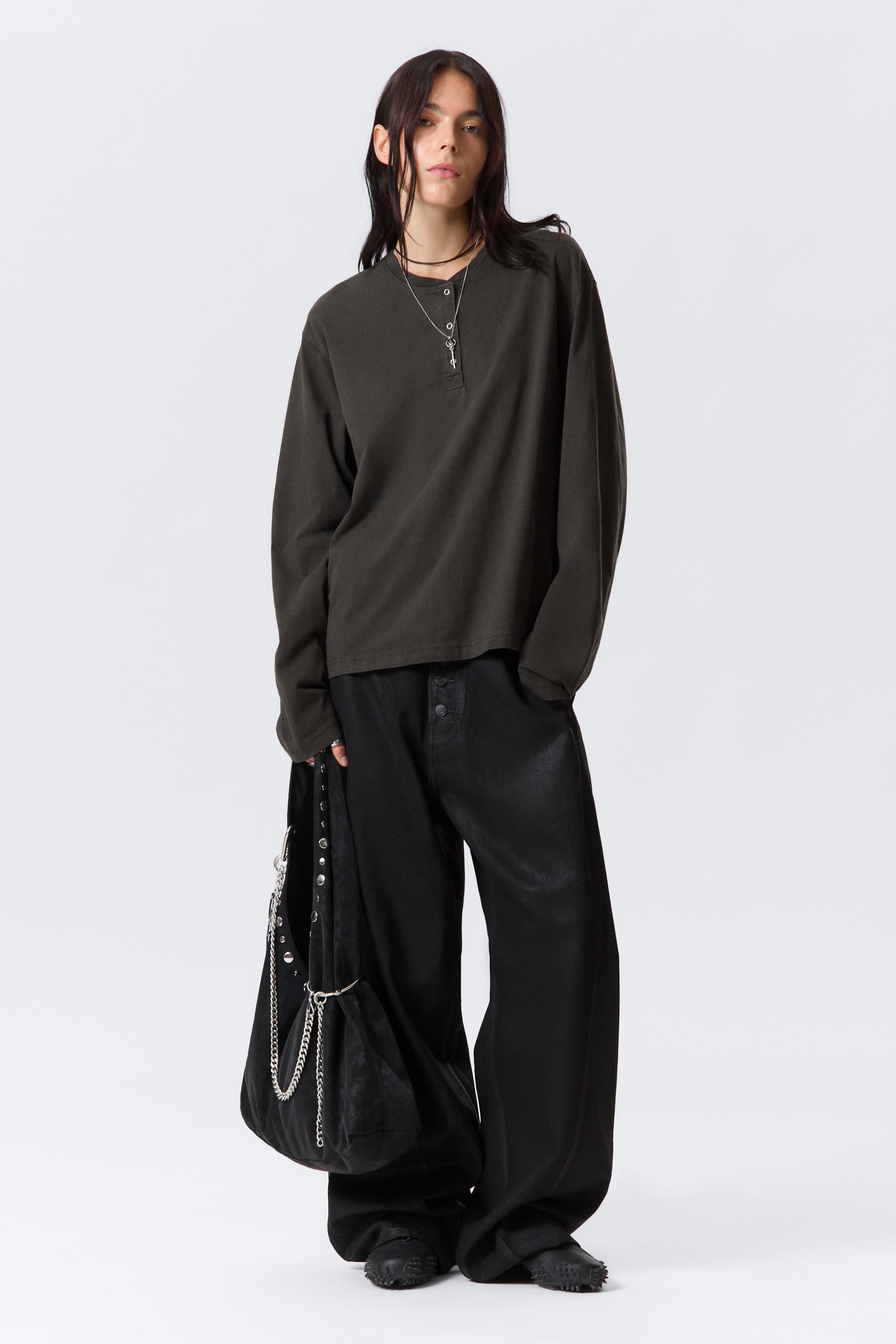 Visa större bild: En mörkgrå långärmad Henley-tröja med en avslappnad, oversized passform presenteras, med en treknappslå vid halsringningen och lösa ärmar. Den bärs med löst sittande svarta byxor med vida ben som har en synlig knapplåsning framtill och en subtil glans, accessoriserad med en svart slapp väska prydd med silvernitar och flera kedjor, samt mörka texturerade platta skor.