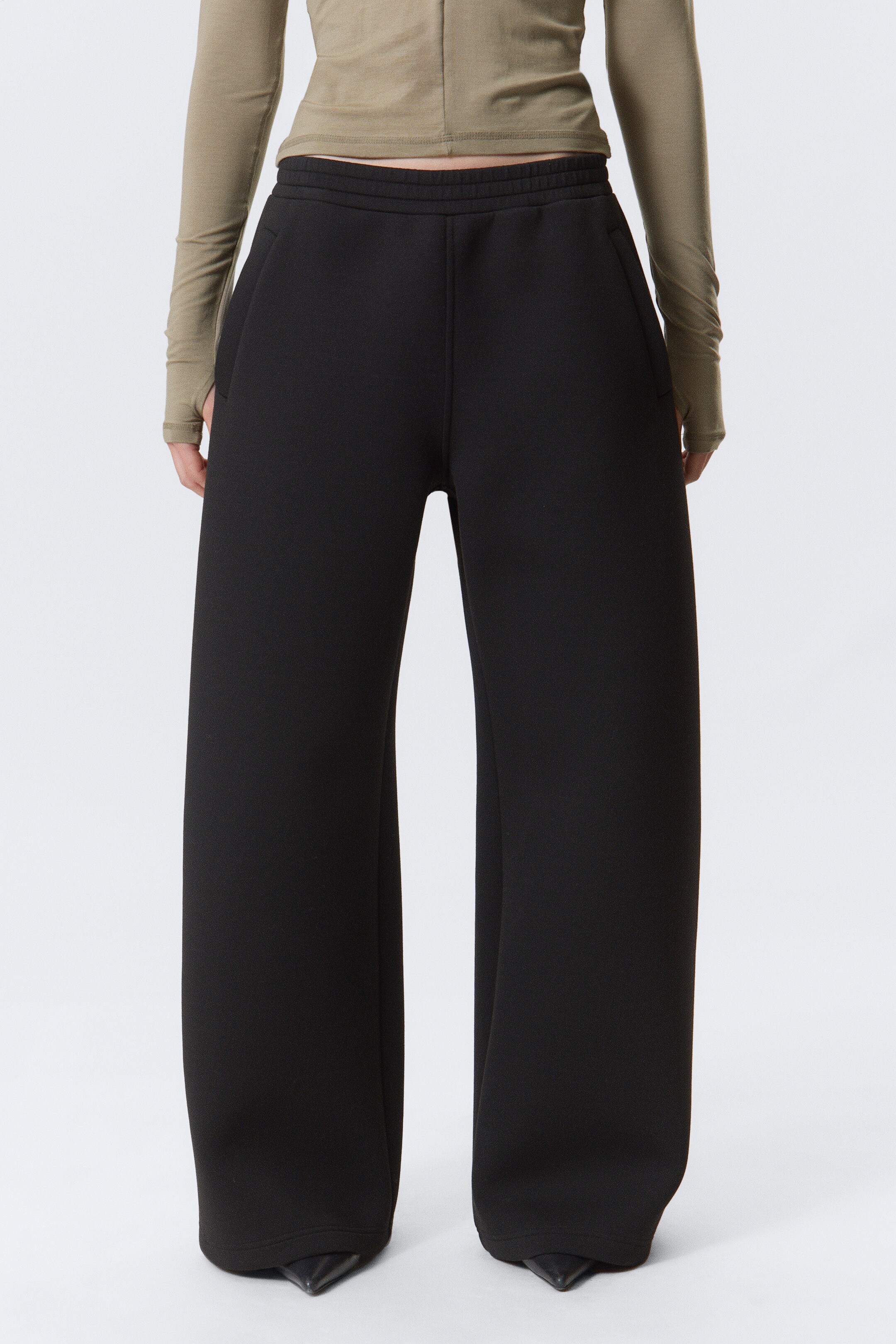Ver imagen más grande: Pantalones negros de pierna ancha, hechos de una tela suave y sustancial, se presentan desde una vista frontal. Presentan una cintura ancha y plana, visible debajo de un top corto color oliva, y bolsillos laterales discretos. Los pantalones muestran un ajuste relajado y fluido, con su longitud completa acumulándose sobre zapatos negros de punta.