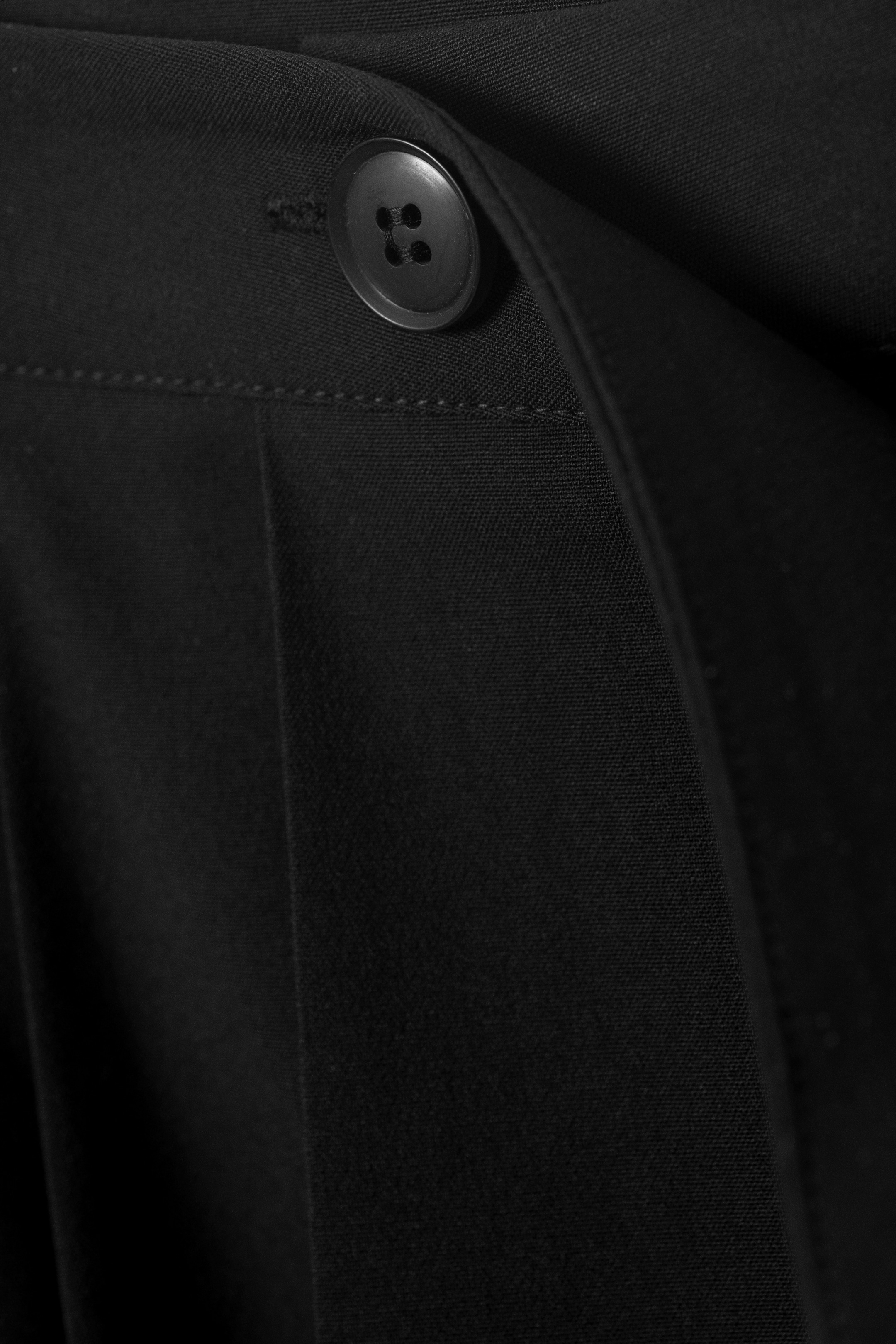 Agrandir l'image: Un vêtement noir montre un gros plan de sa fermeture avant, où un bouton noir à quatre trous fixe le tissu superposé. Une couture horizontale est visible au-dessus du bouton, et un pli vertical se forme dans le tissu en dessous.
