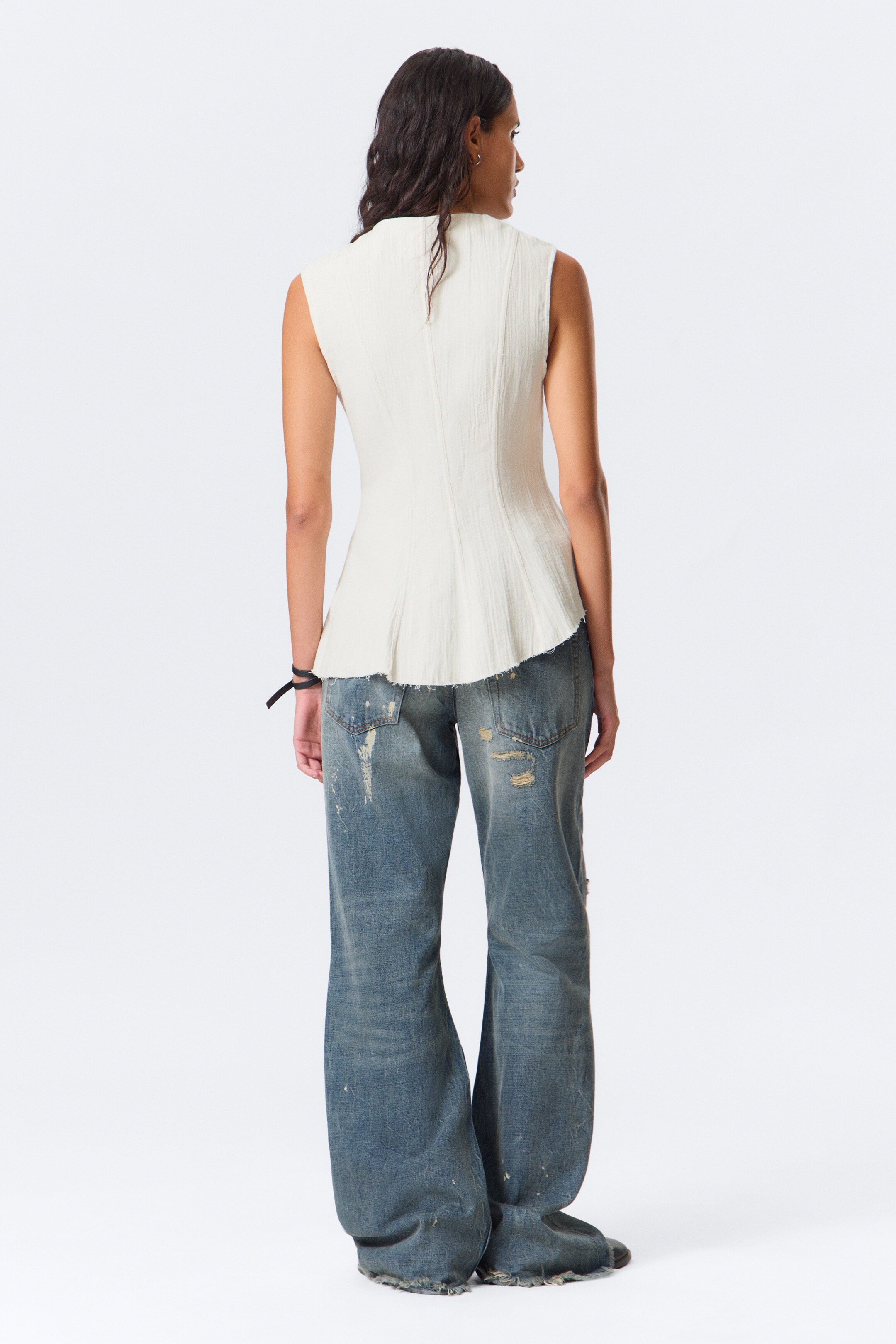 Vis større bilde: En off-white teksturert ermeløs topp med høy halsutringning og en ujevn råkantet peplumfald vises fra baksiden, stylet med distressed blå vide jeans med rifter og malingsflekker.