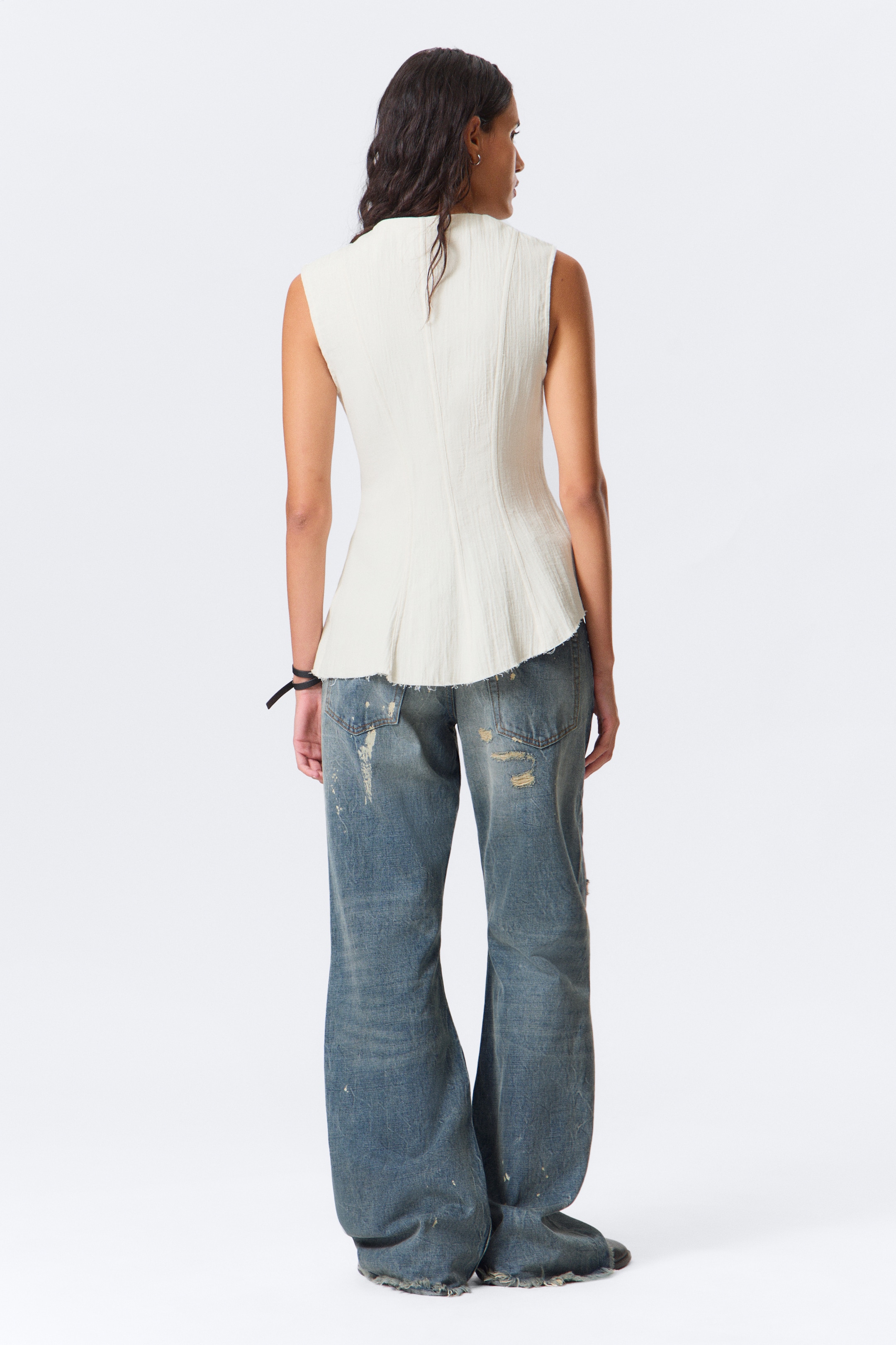 Grotere afbeelding bekijken: Een off-white getextureerde mouwloze top met een hoge halslijn en een ongelijke, rauwe peplumzoom wordt van de achterkant getoond, gestyled met blauwe wijde jeans met scheuren en verfspatten.