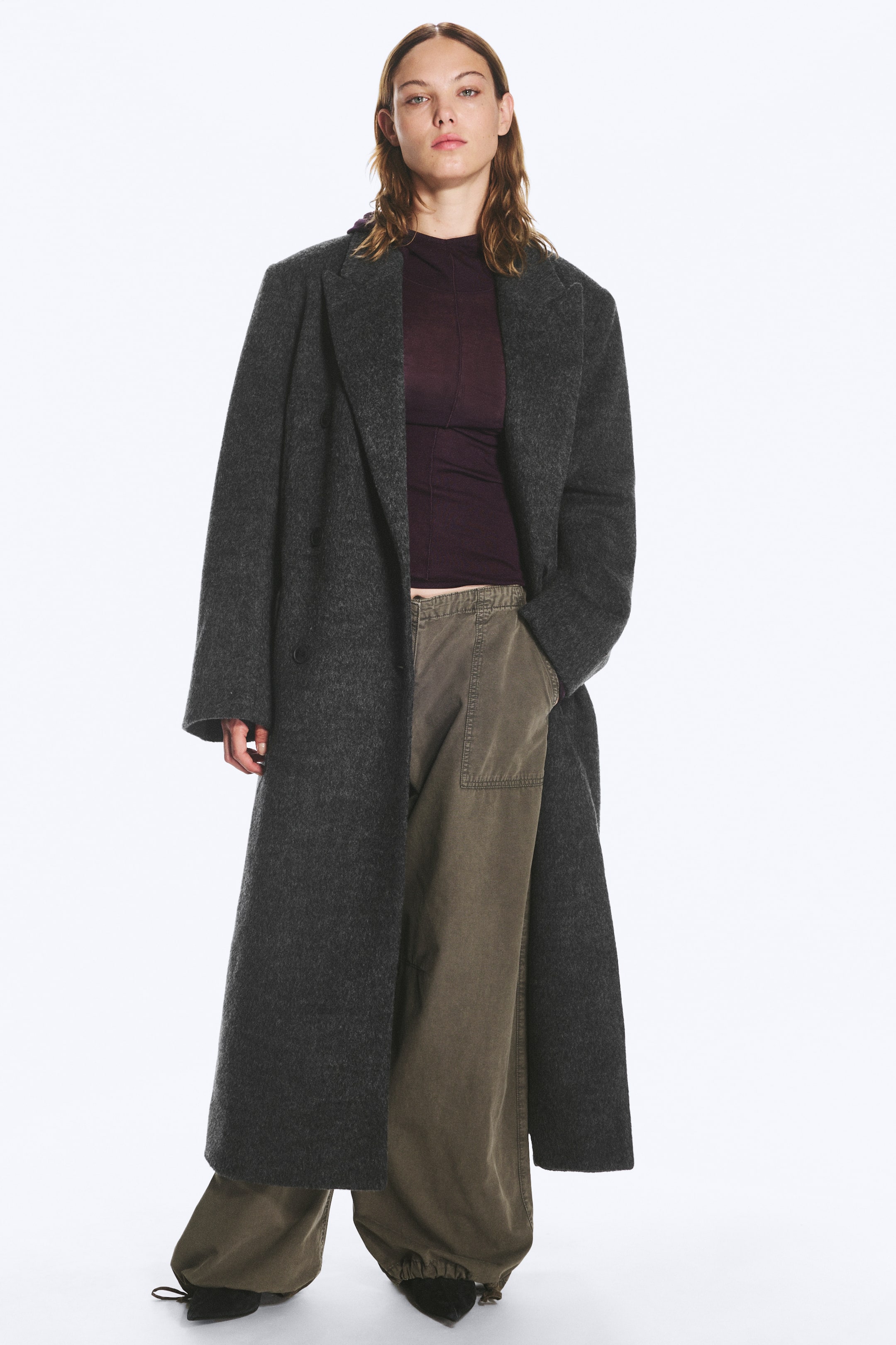 Visualizza immagine più grande: Un cappotto oversize in misto lana grigio scuro con ampio colletto a revers viene indossato aperto dalla modella. Il cappotto lungo è abbinato a un top viola scuro a collo alto e a pantaloni verdi a gamba larga.