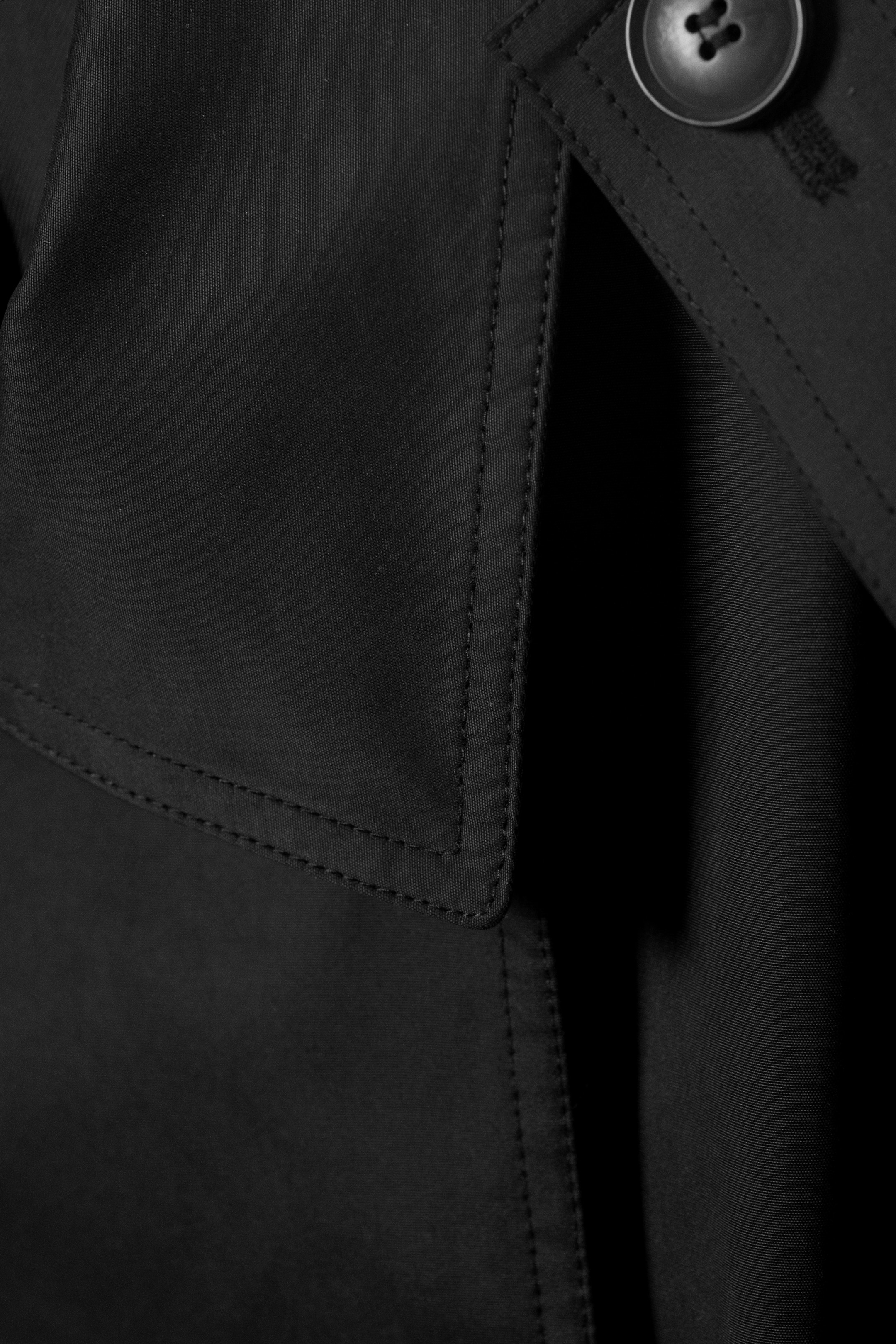 Visualizza immagine più grande: Un trench coat nero viene mostrato in primo piano, mettendo in risalto il tessuto strutturato della sua pattina antipioggia, che presenta un bottone prominente e cuciture visibili.
