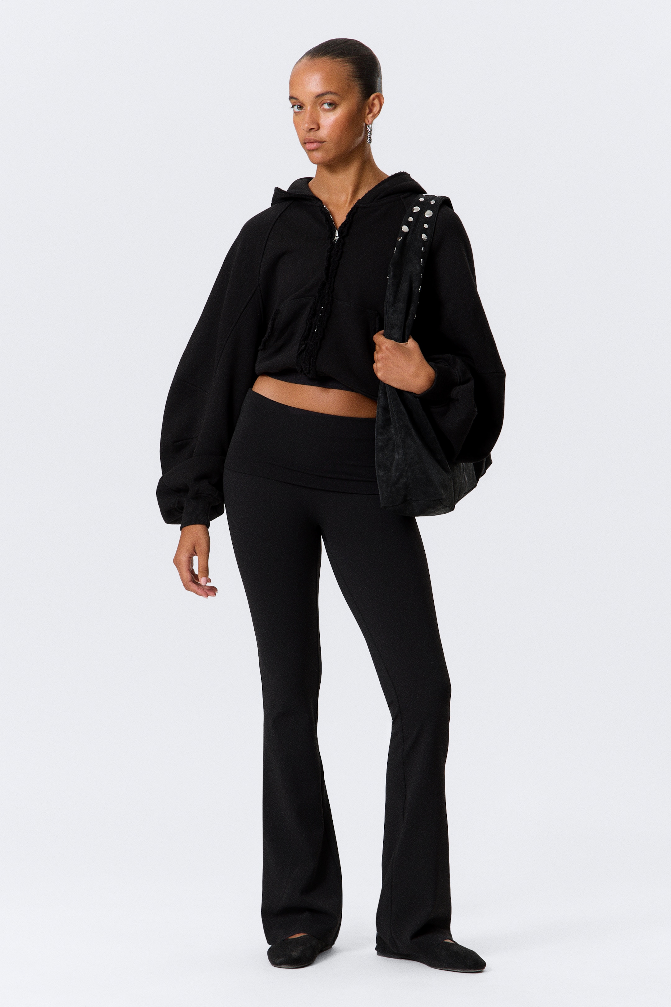 Visa större bild: En svart kort hoodie, med en dragkedja framtill med slitna detaljer, en fastsydd huva och voluminösa ärmar, presenteras tillsammans med svarta högmidjade utsvängda leggings, som är åtsittande genom låret och utsvängda från knät. En svart axelväska med silverfärgade nitar bärs på axeln.